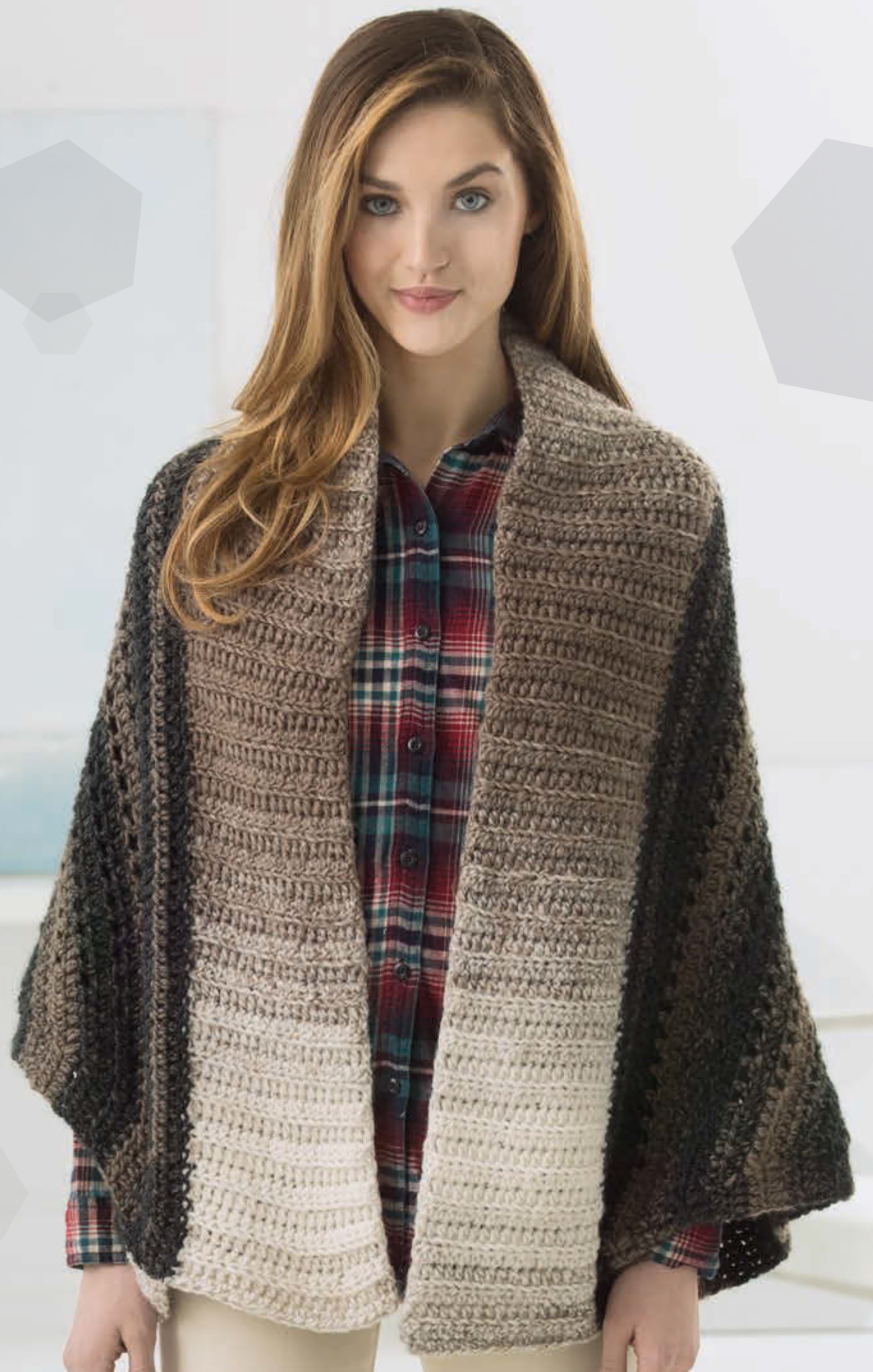 A woman with long brown hair wears a plaid shirt and a crocheted shawl made with Leisure Arts Simply Scarfie Crochet yarn in gradient brown to cream, standing indoors against a light background.