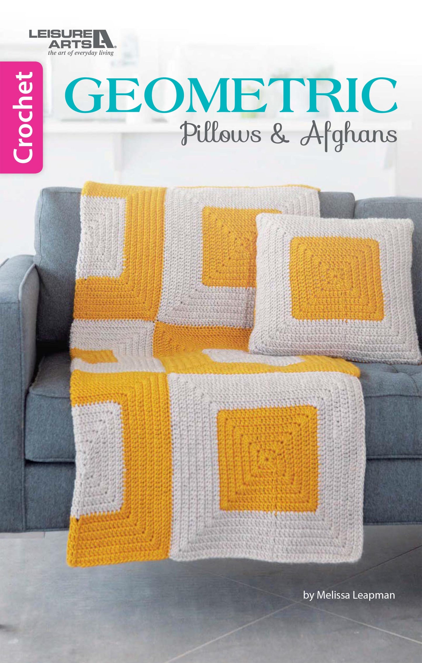 A gray couch features a crocheted afghan and two pillows with white, gray, and yellow geometric squares. The book "Geometric Pillows & Afghans" by Leisure Arts is perfect for beginners to explore new crochet patterns.