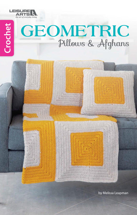 A gray couch features a crocheted afghan and two pillows with white, gray, and yellow geometric squares. The book "Geometric Pillows & Afghans" by Leisure Arts is perfect for beginners to explore new crochet patterns.
