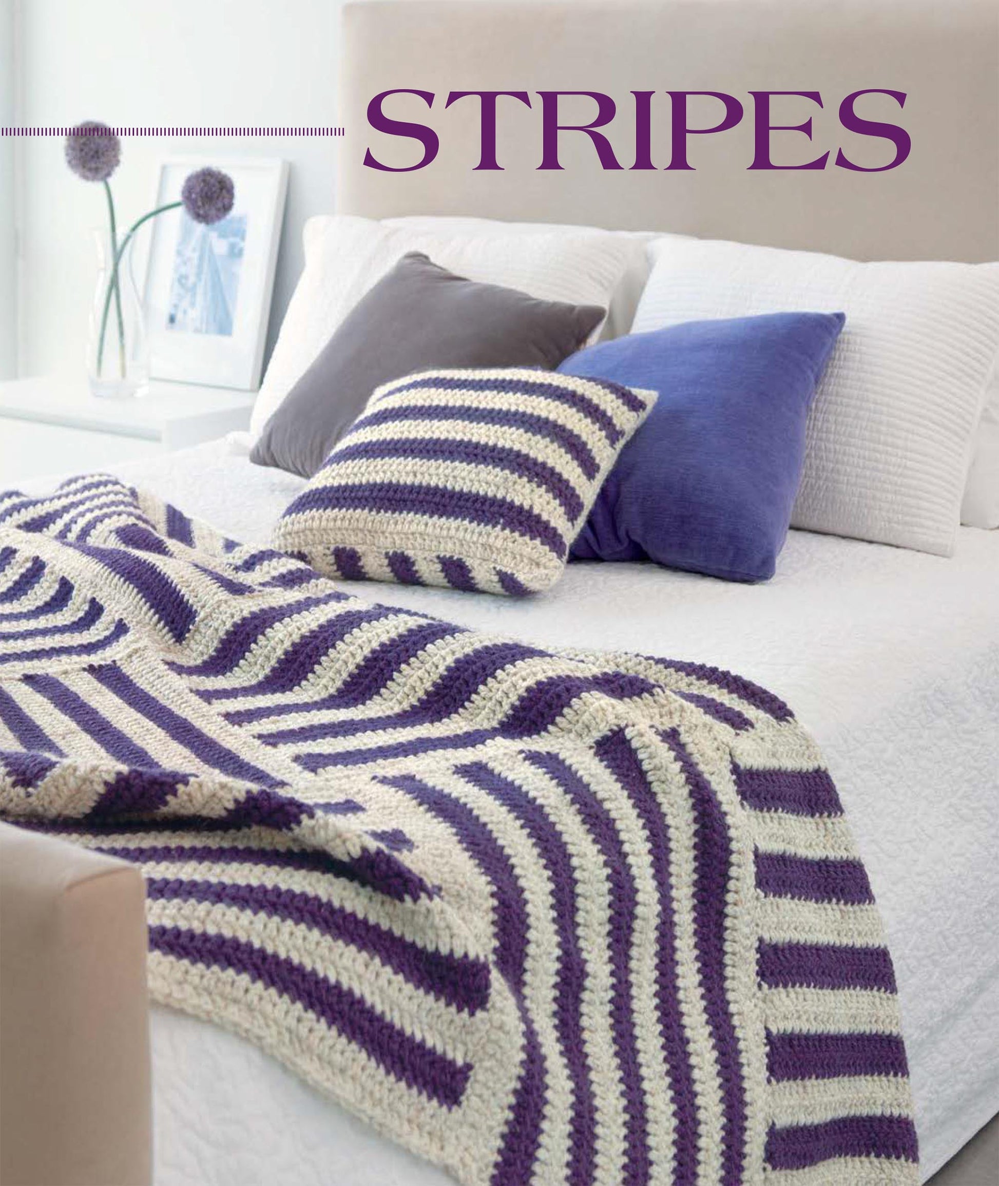 Showcase your beginner crochet skills or new patterns with the Geometric Pillows & Afghans from Leisure Arts, featuring purple pillows, white linens, and a striped crocheted blanket and pillow. Decorative pieces and a framed photo accent the nightstand.