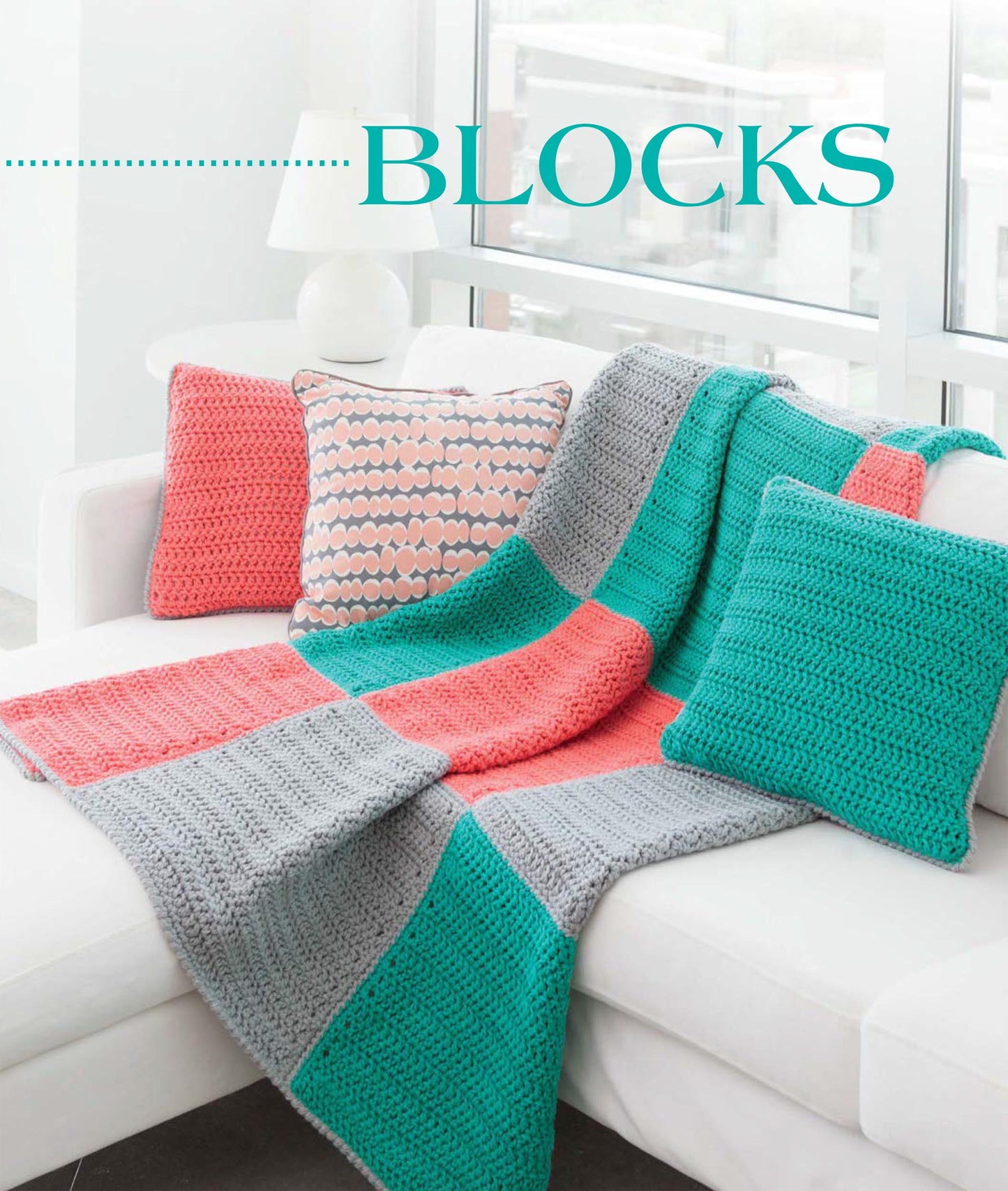 The white sofa features Geometric Pillows & Afghans by Leisure Arts—a crocheted blanket in teal, gray, and coral blocks with three matching pillows—ideal for showcasing beginner crochet or exploring new patterns. Lamp and window appear in the background.