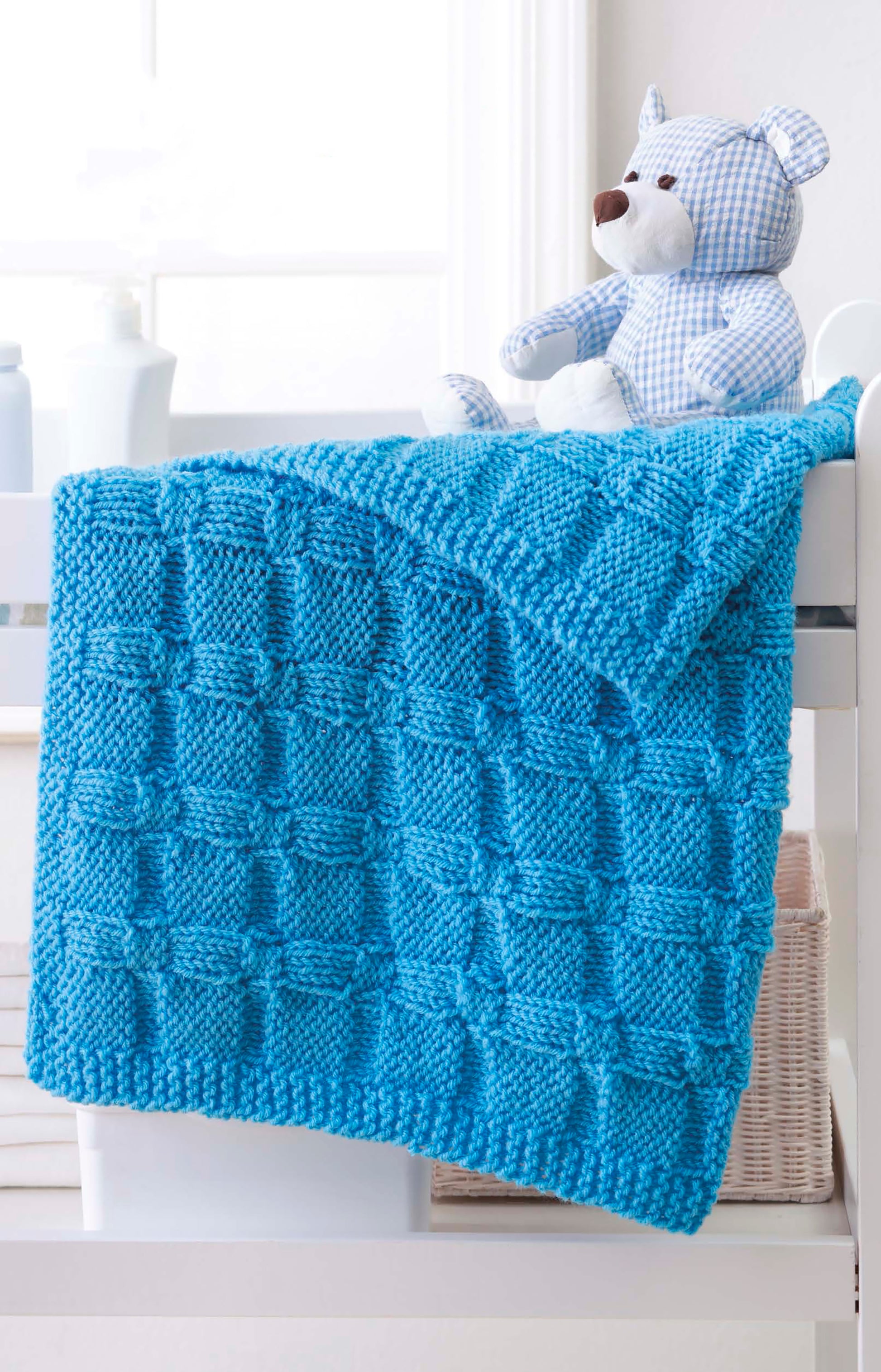 A vibrant blue baby blanket, made using Leisure Arts' "Make in a Weekend Afghans to Knit," is draped over a white crib rail with a blue and white gingham teddy bear beside it.