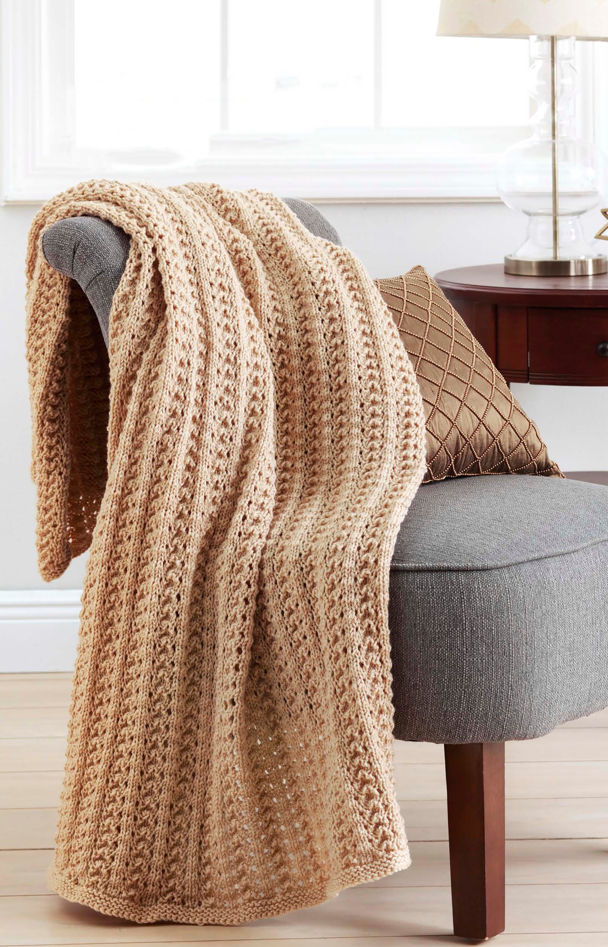 A beige knitted blanket from Leisure Arts’ "Make in a Weekend Afghans to Knit" drapes over a gray chair next to a dark wood side table with a brown pillow and glass lamp, creating a bright and cozy room.