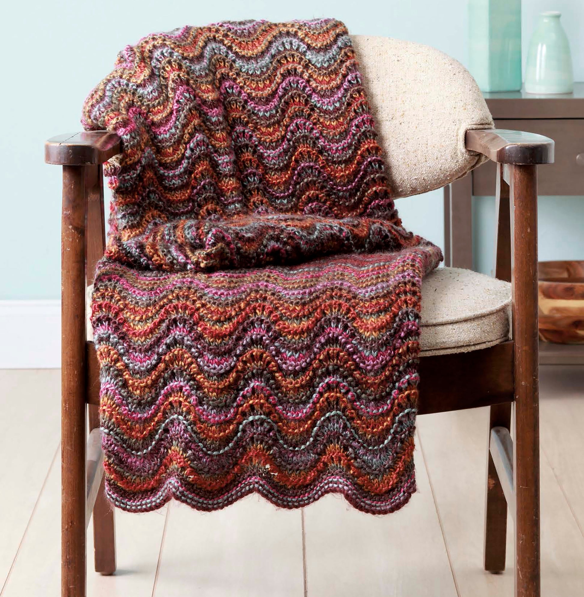 The "Make in a Weekend Afghans to Knit" book by Leisure Arts offers beginner-friendly patterns, like the multicolored wavy knit blanket shown draped over a beige-cushioned wooden chair, with stylish decor in the background.
