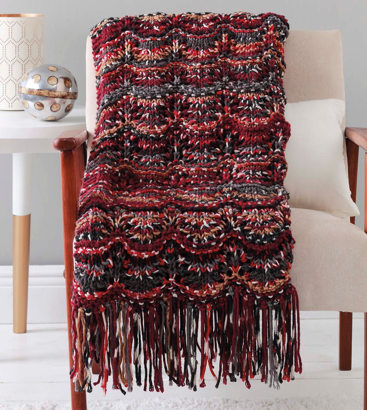 A red, black, and brown crocheted blanket with a scalloped pattern and fringe draped over a beige armchair showcases cozy style—great inspiration from "Make in a Weekend Afghans to Knit" by Leisure Arts for quick, beginner projects.