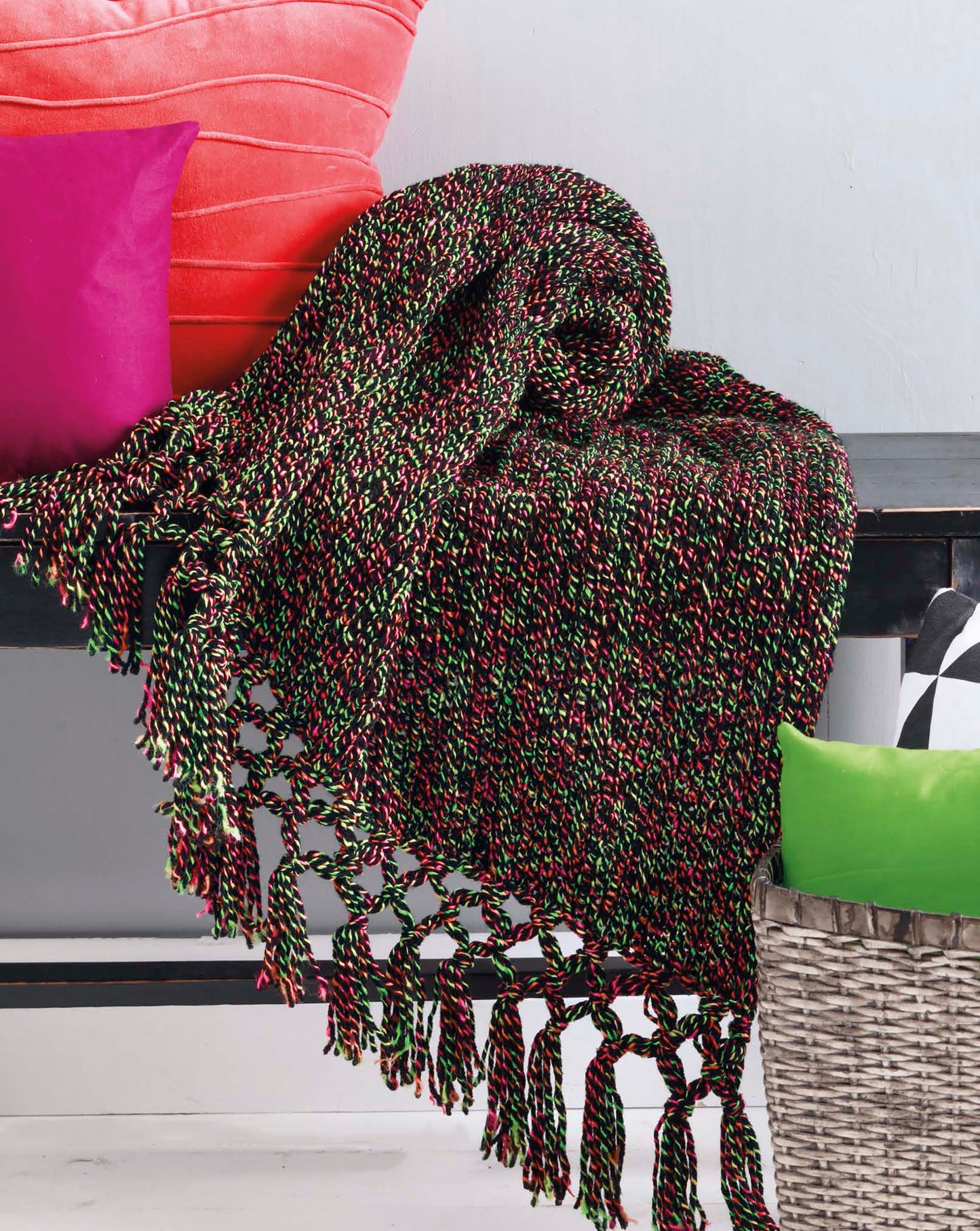 A multicolored woven throw from the Leisure Arts "Make in a Weekend Afghans to Knit" collection drapes over a dark bench with bright pink, green, and black-and-white pillows. A gray basket sits beside the bench.