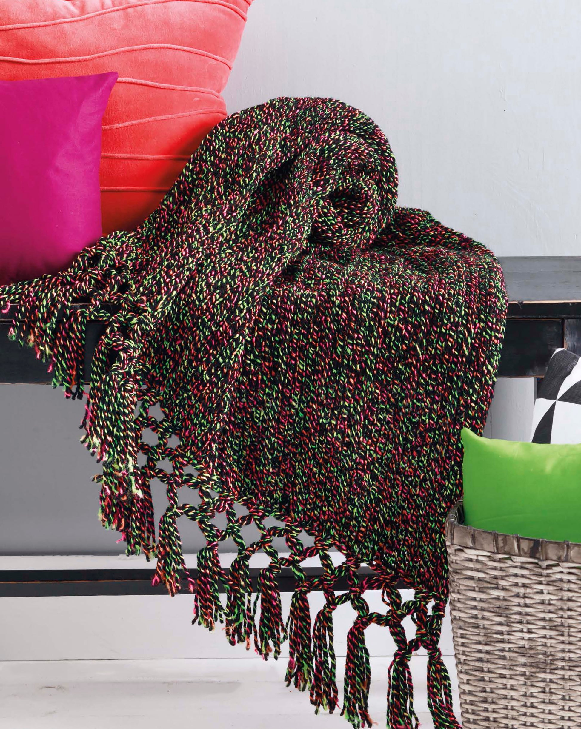 A multicolored woven throw from the Leisure Arts "Make in a Weekend Afghans to Knit" collection drapes over a dark bench with bright pink, green, and black-and-white pillows. A gray basket sits beside the bench.