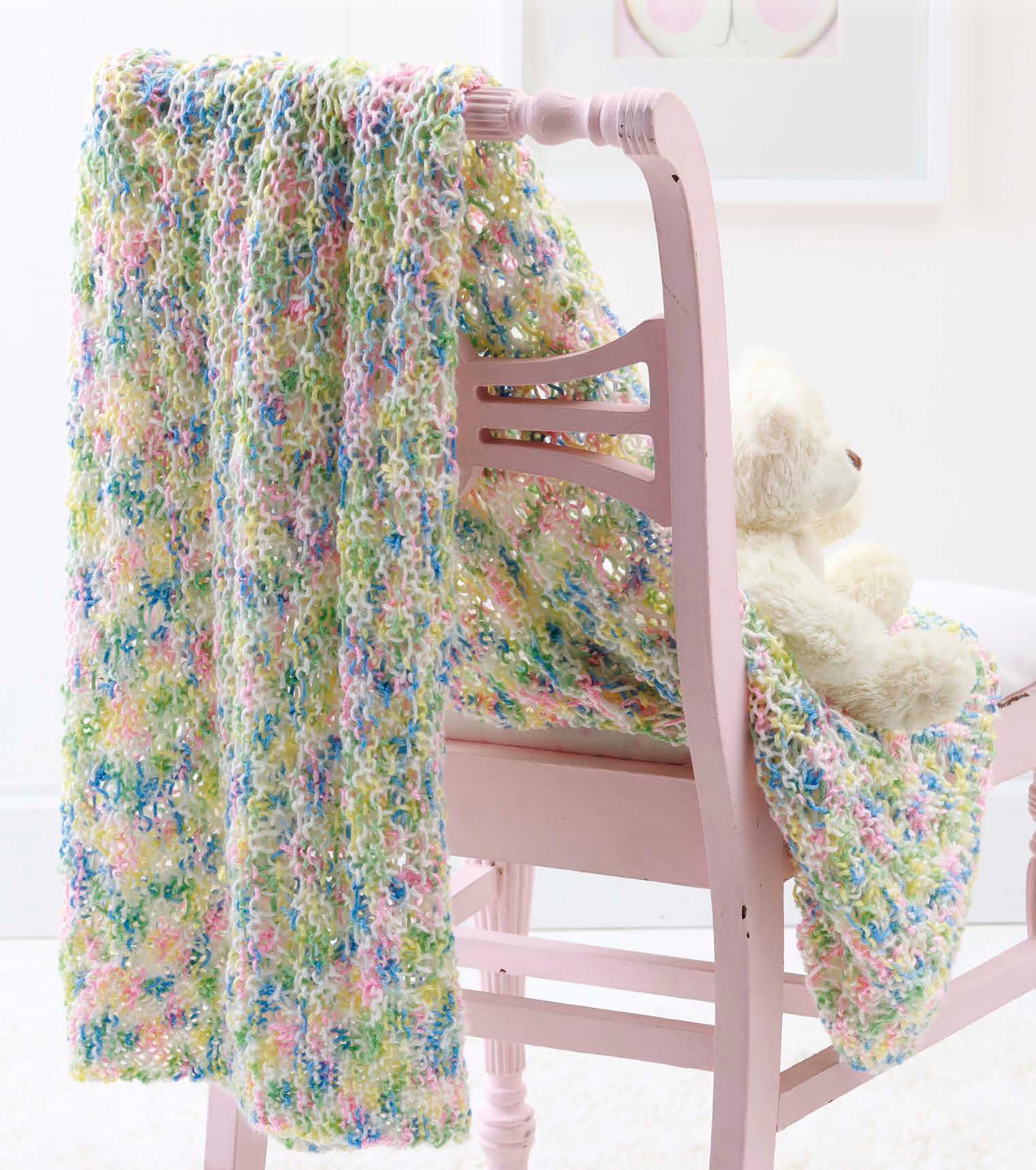 A pastel multicolored blanket from the Make in a Weekend Afghans to Knit book by Leisure Arts is draped over a light pink wooden chair, with a white teddy bear on the seat and soft, light decor in the background.