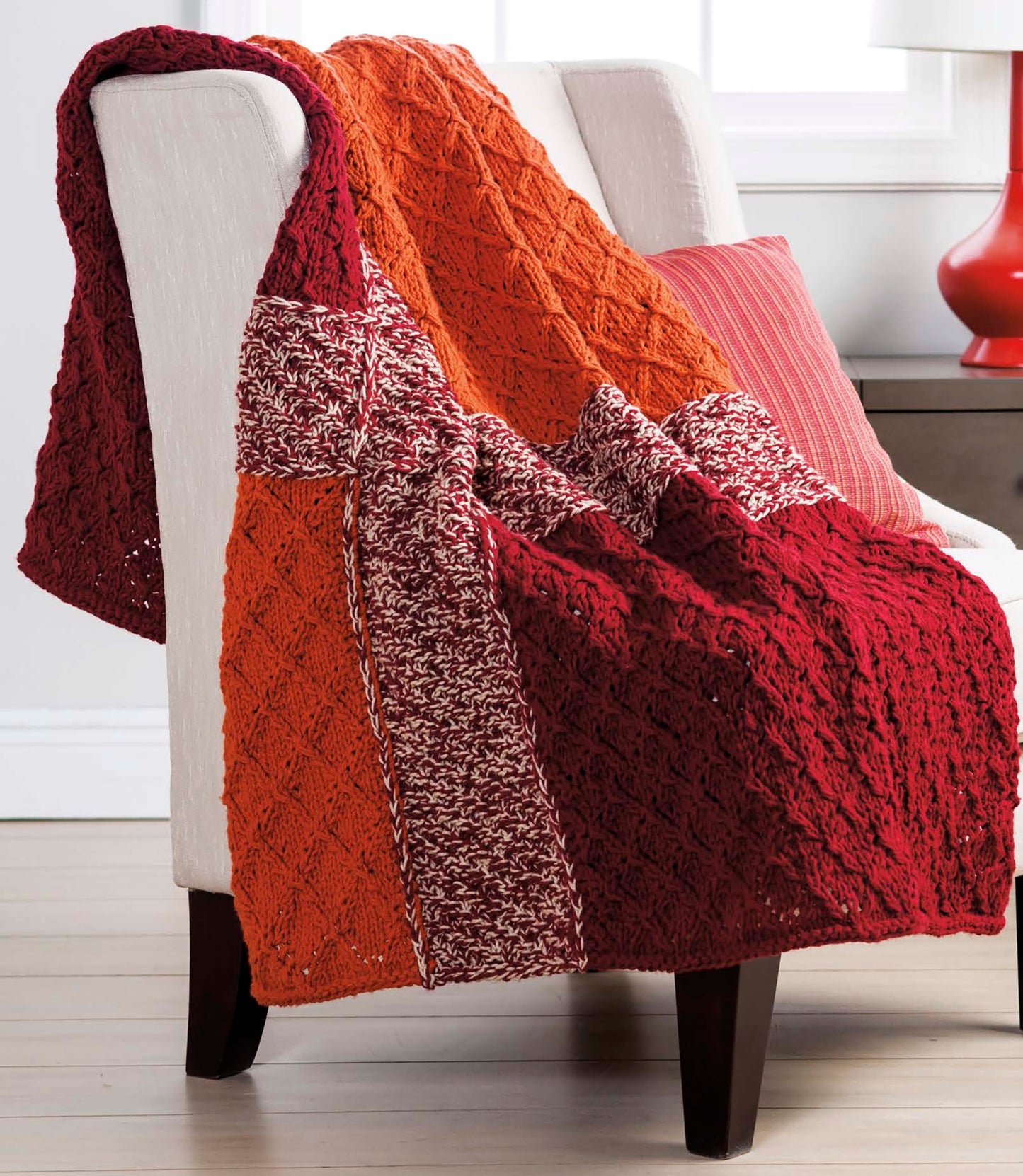 A white armchair features a textured knit blanket in red, orange, and maroon from Leisure Arts’ Quick Throws to Knit, perfect for fans of easy patterns. It's paired with a striped red pillow and a red lamp on a small table in the background.
