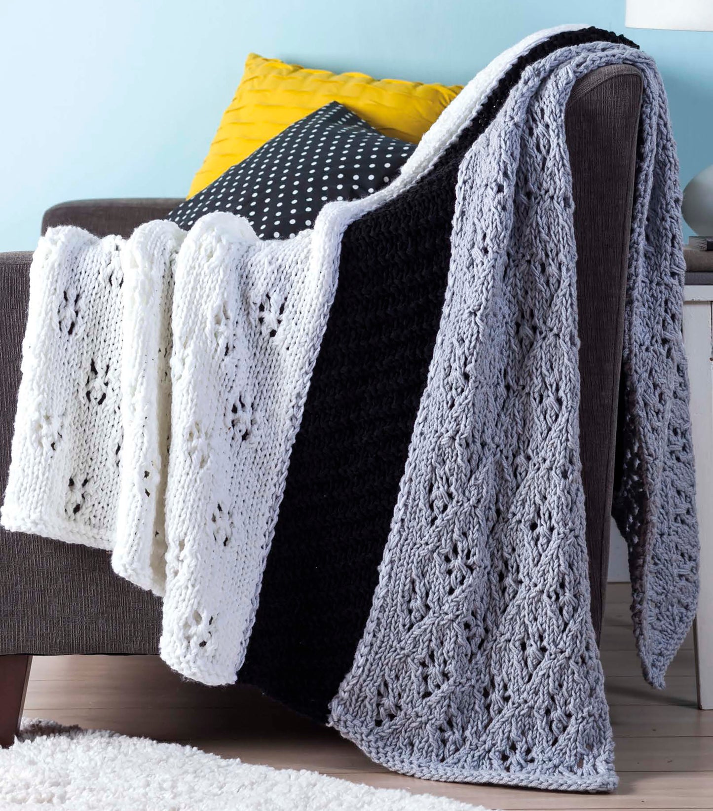 The "Quick Throws to Knit" by Leisure Arts features a chunky knitted blanket with white, gray, and black panels draped over a gray armchair, complemented by yellow and black polka dot pillows in a modern living room.