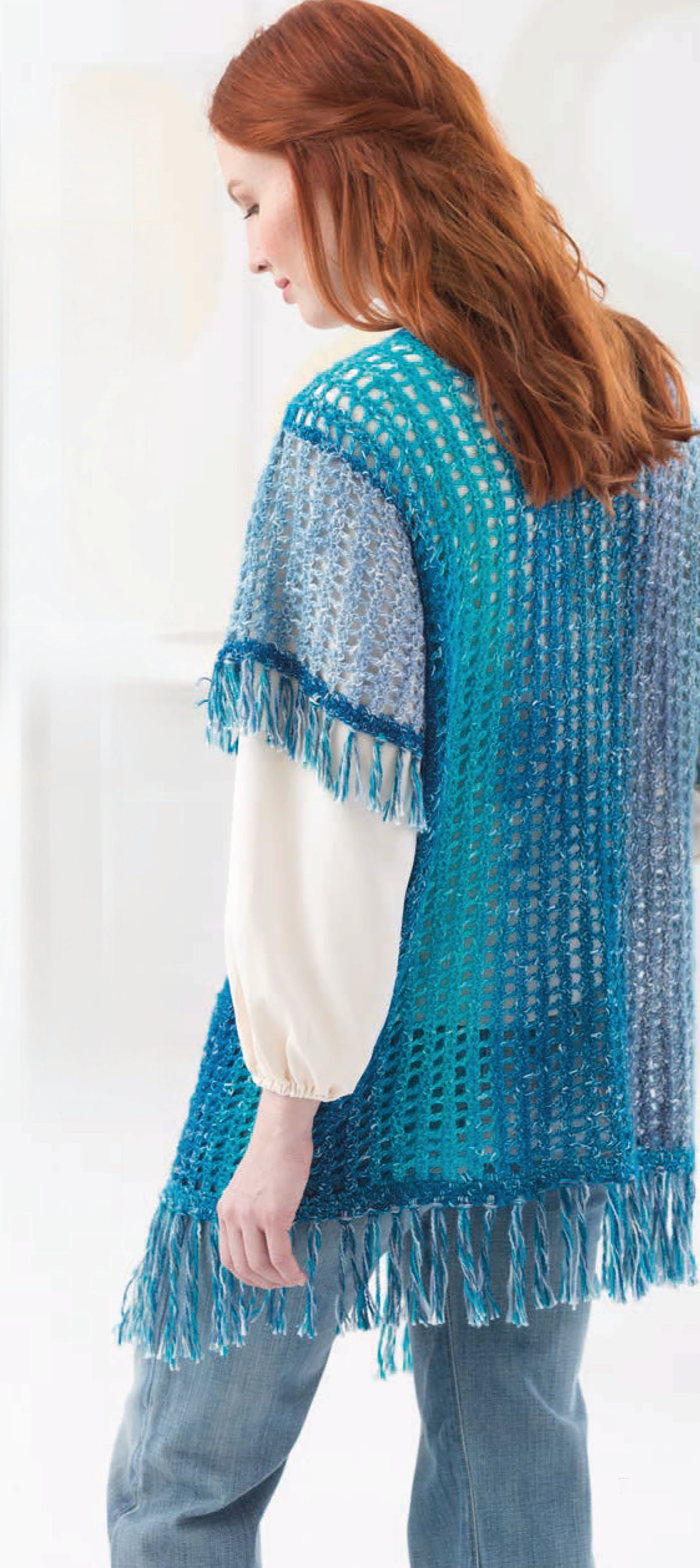 A woman with long red hair models a blue and turquoise fringed poncho from Leisure Arts’ “Shawls & Vests—Crochet 7 Bohemian Designs,” worn over a white blouse and jeans, standing indoors, facing to the side.