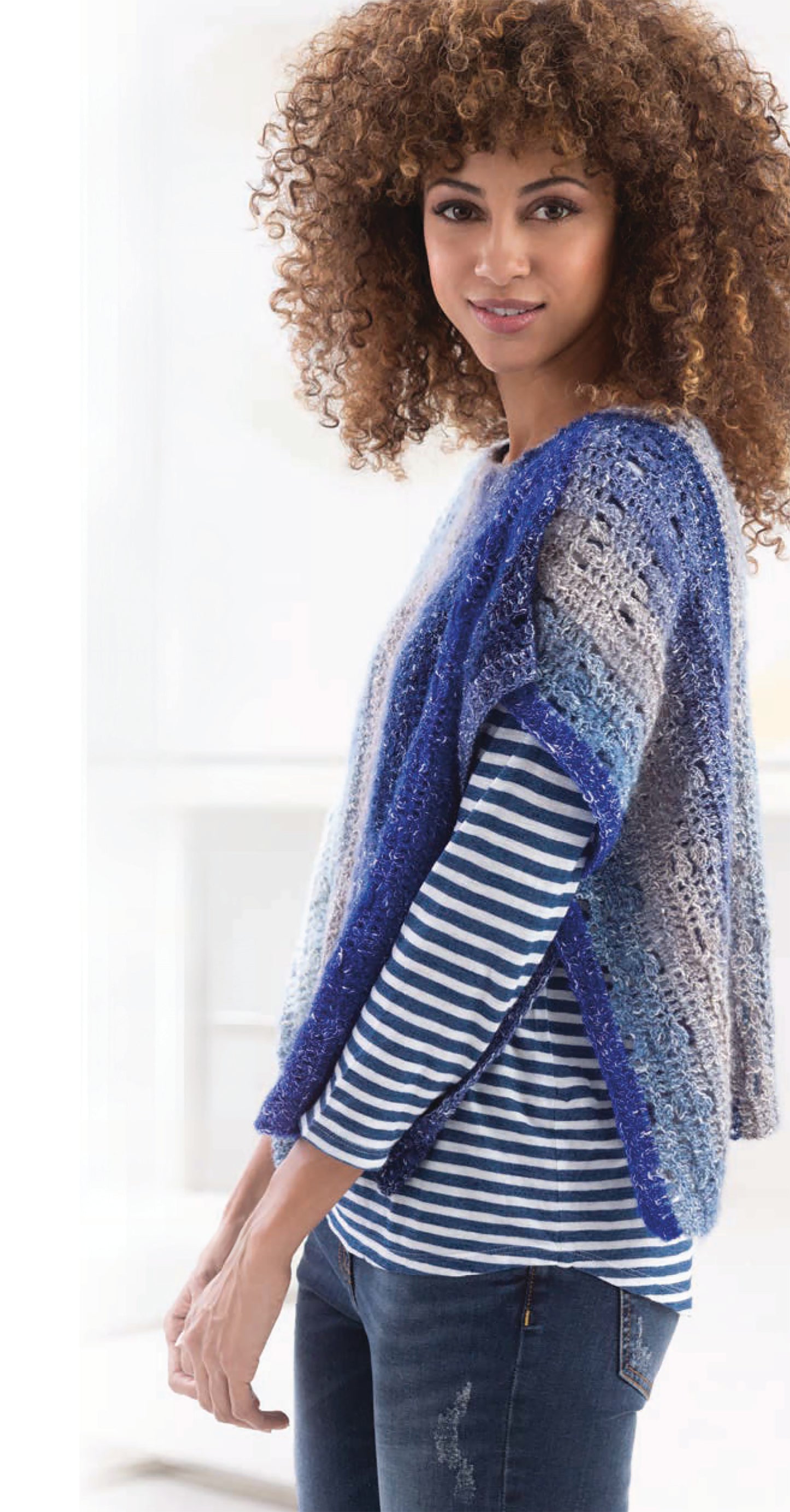 A woman with curly hair wears a blue and gray poncho from Leisure Arts' "Shawls & Vests—Crochet 7 Bohemian Designs" over a navy and white striped shirt and blue jeans, standing indoors in natural light.