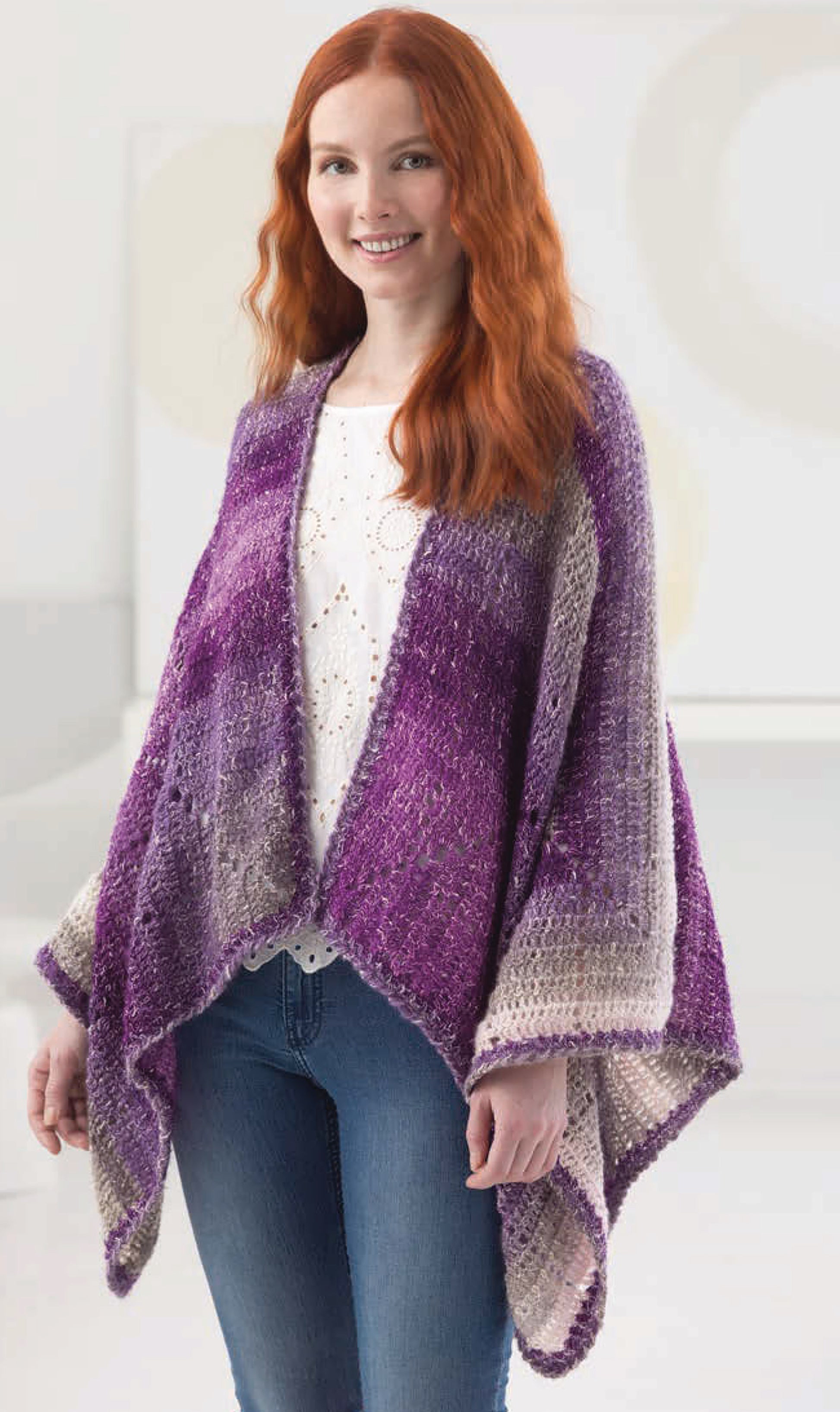 A woman with long red hair models a purple and lavender shawl from "Shawls & Vests—Crochet 7 Bohemian Designs" by Leisure Arts, wearing a white top and blue jeans, smiling indoors and highlighting trendy crochet patterns.