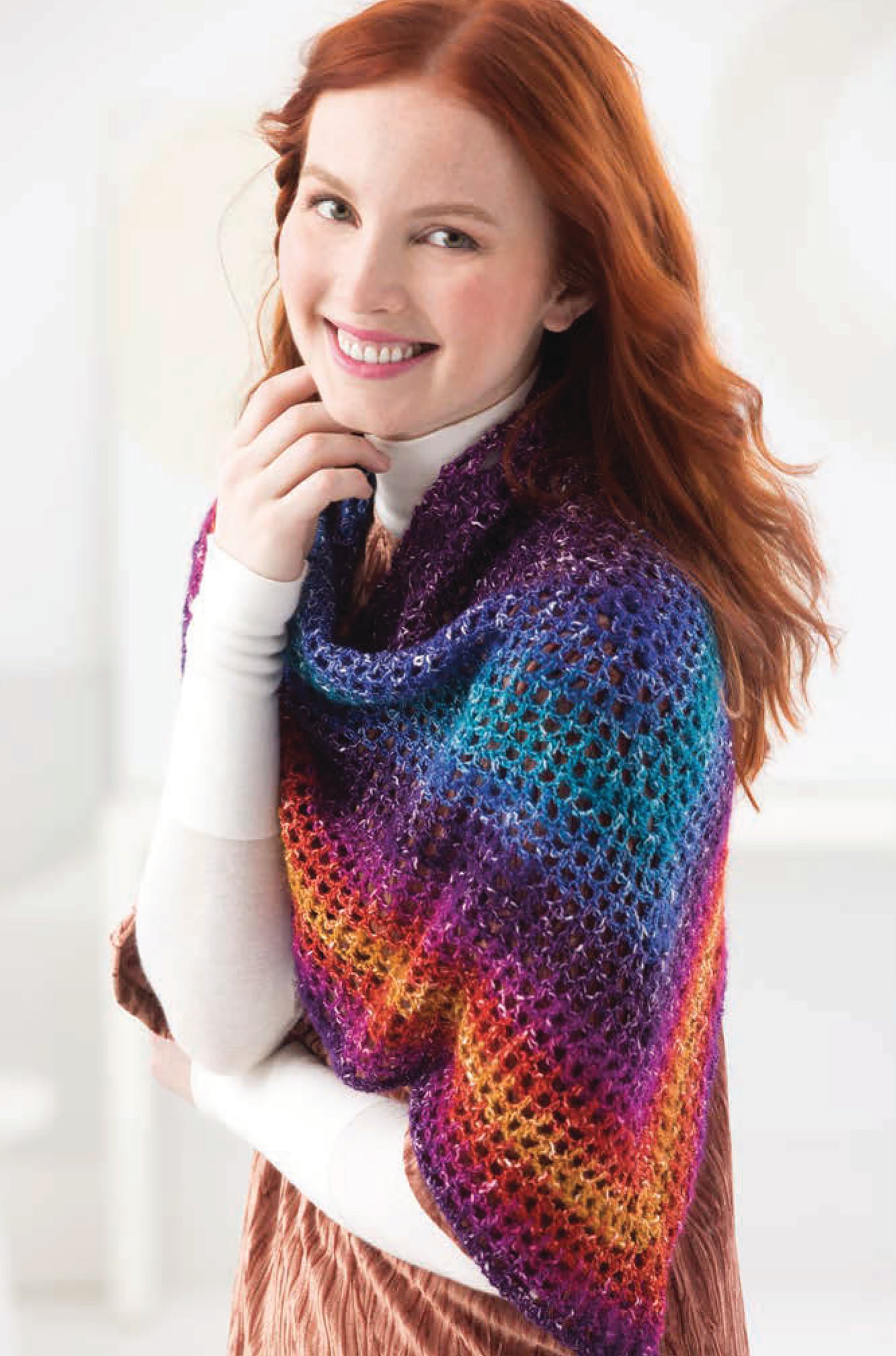A woman with long red hair smiles while modeling a colorful lacy shawl with purple, blue, pink, orange, and yellow stripes from "Shawls & Vests—Crochet 7 Bohemian Designs" by Leisure Arts over a white turtleneck and light brown dress.