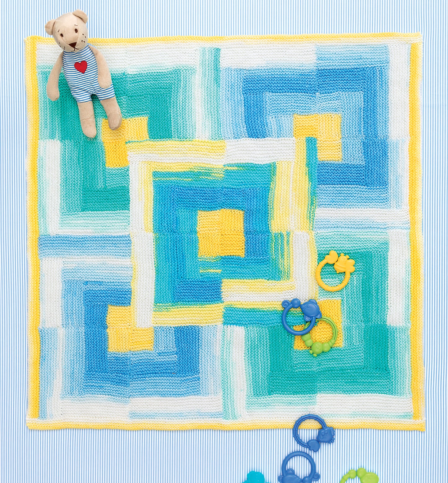 The Ice Cream Baby Afghans Digital Download by Leisure Arts features a colorful geometric knit blanket in blue, yellow, green, and white. Displayed on a striped surface with a teddy bear and blue and yellow baby rattles.