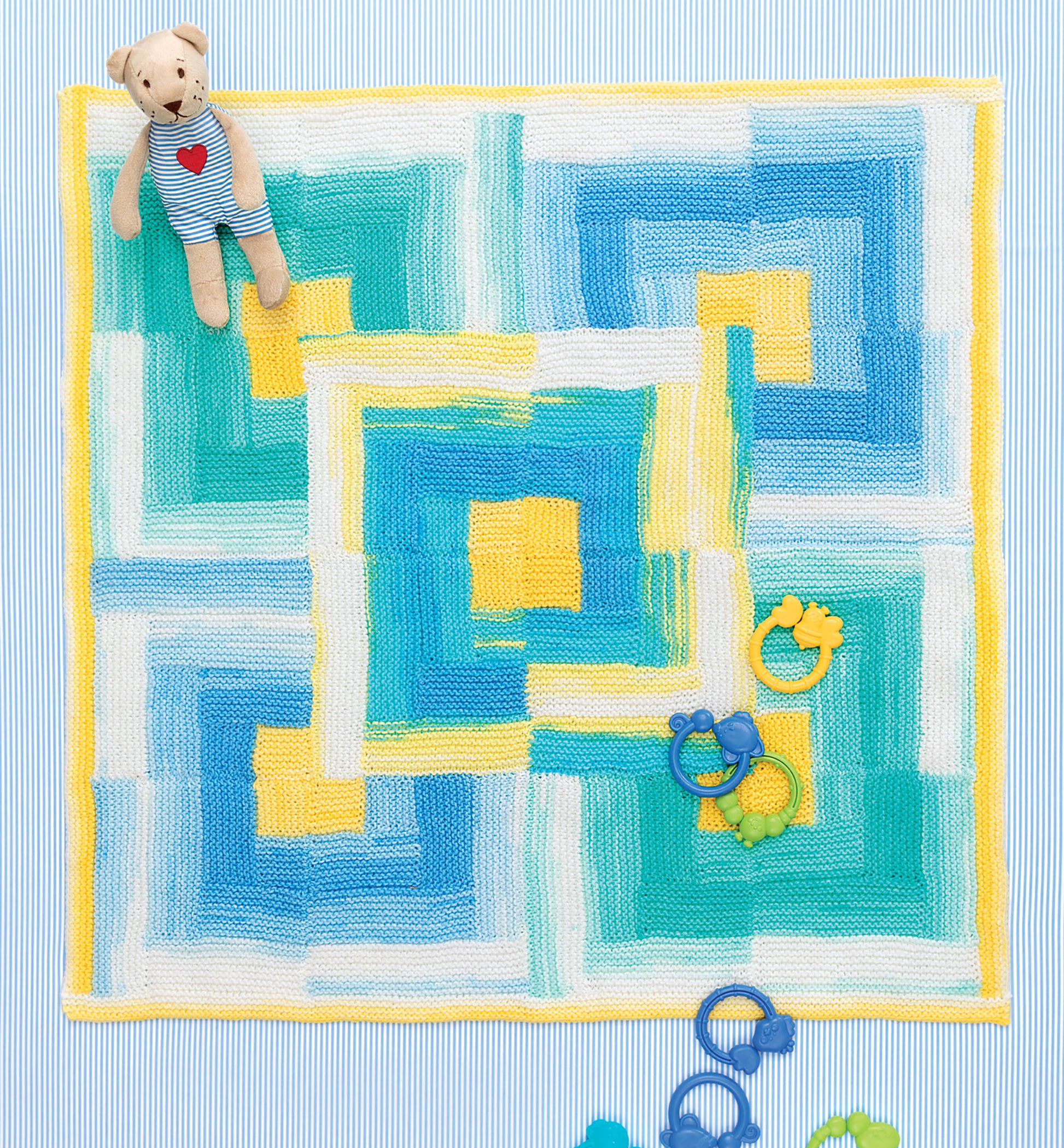 The Ice Cream Baby Afghans Digital Download by Leisure Arts features a colorful geometric knit blanket in blue, yellow, green, and white. Displayed on a striped surface with a teddy bear and blue and yellow baby rattles.
