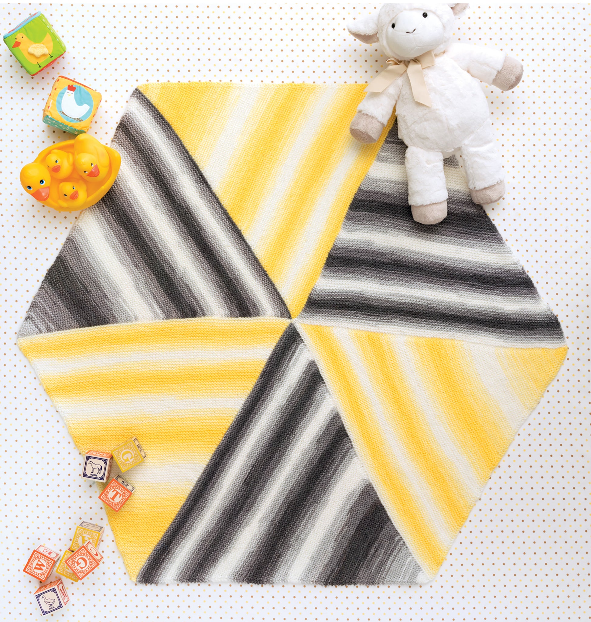 The Ice Cream Baby Afghans Digital Download by Leisure Arts features a hexagonal baby mat in self-striping yellow, black, gray, and white yarn, shown on a dotted surface with toy blocks, rubber ducks in a dish, and a stuffed lamb.