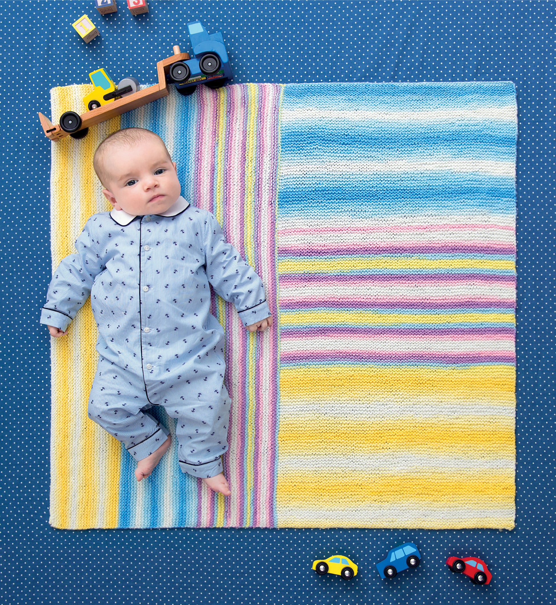 A baby in a blue onesie lies on a colorful striped blanket made with Leisure Arts' Ice Cream Baby Afghans Digital Download, surrounded by toy cars and trucks on a blue polka-dot background, gazing upward as toys are arranged around their head and feet.