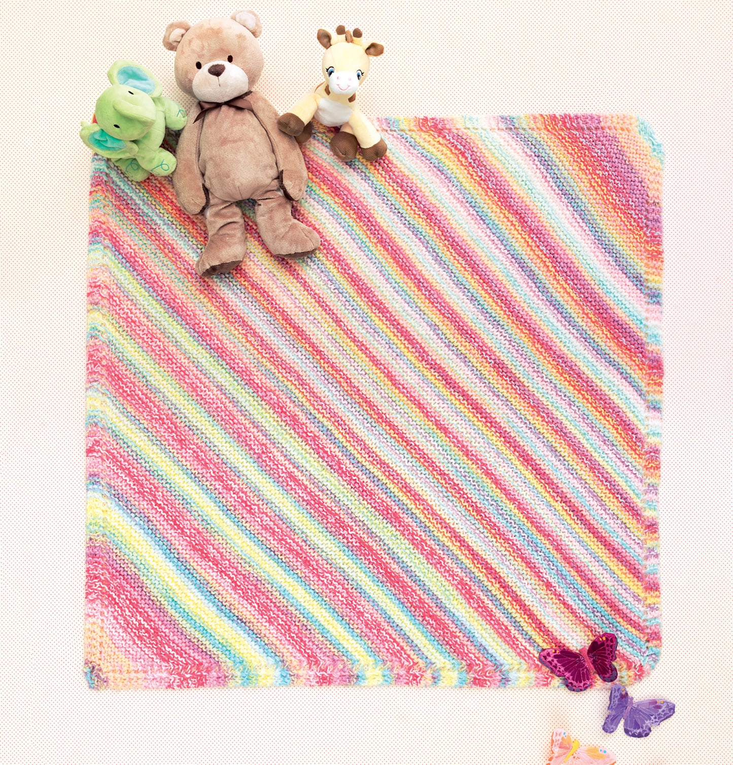 The Ice Cream Baby Afghans Digital Download by Leisure Arts features a pastel rainbow-striped knitted baby blanket spread out flat with three stuffed animals in one corner and two purple butterflies in the opposite corner.