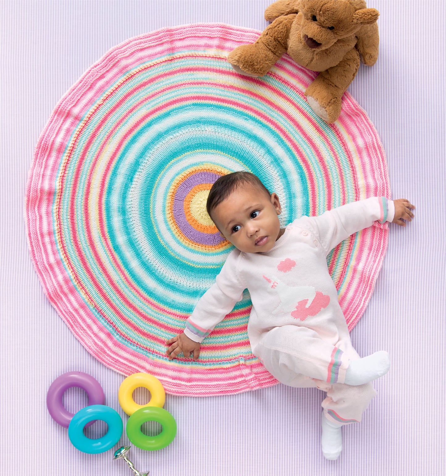 A baby in a pink and white onesie lies on a colorful mat made with self-striping yarn, crafted from the Ice Cream Baby Afghans Digital Download by Leisure Arts. A teddy bear and three ring-shaped toys in purple, turquoise, and yellow are nearby.