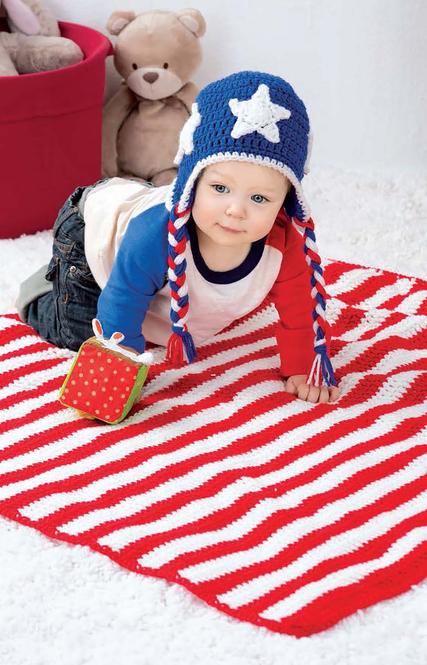 A baby from the Baby Collection—by Leisure Arts wears a blue hat with a white star and red-and-white braids, crawling on a crochet blanket with a soft toy block. A stuffed bear adds charm to this adorable crochet scene.