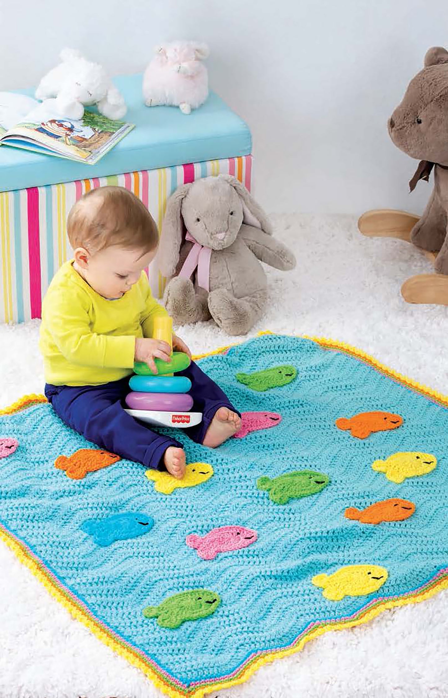 A baby sits on a blue crochet blanket from the Baby Collection by Leisure Arts, crafted with beginner patterns and decorated with colorful fish and toy rings. Plush animals, a striped bench, and books are in the background on a soft white carpet.