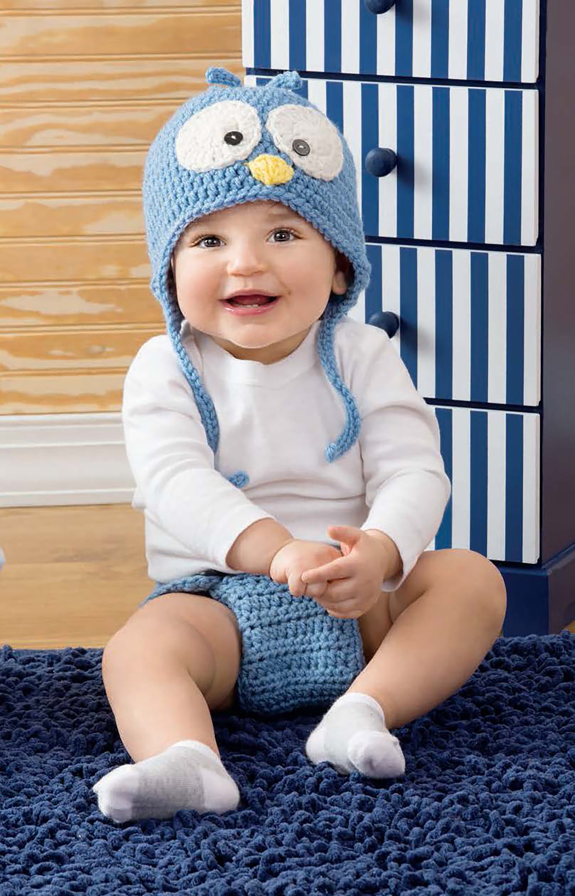 A smiling baby models items from Leisure Arts’ Baby Collection—blue crocheted owl hat, white long-sleeve shirt, blue diaper cover—sitting on a dark blue rug before a blue and white striped dresser.