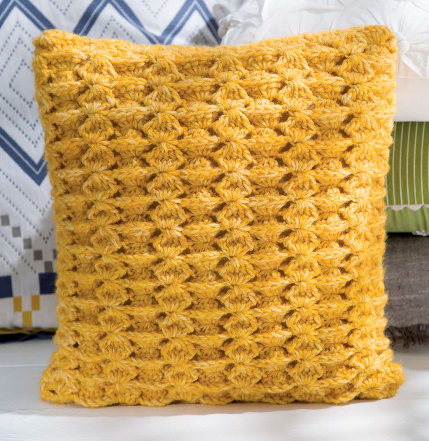 The Leisure Arts Textured Pillows Crochet features a yellow pillow with a shell-like pattern in front of geometric and striped designs, adding unique textured crochet style and depth to any space.
