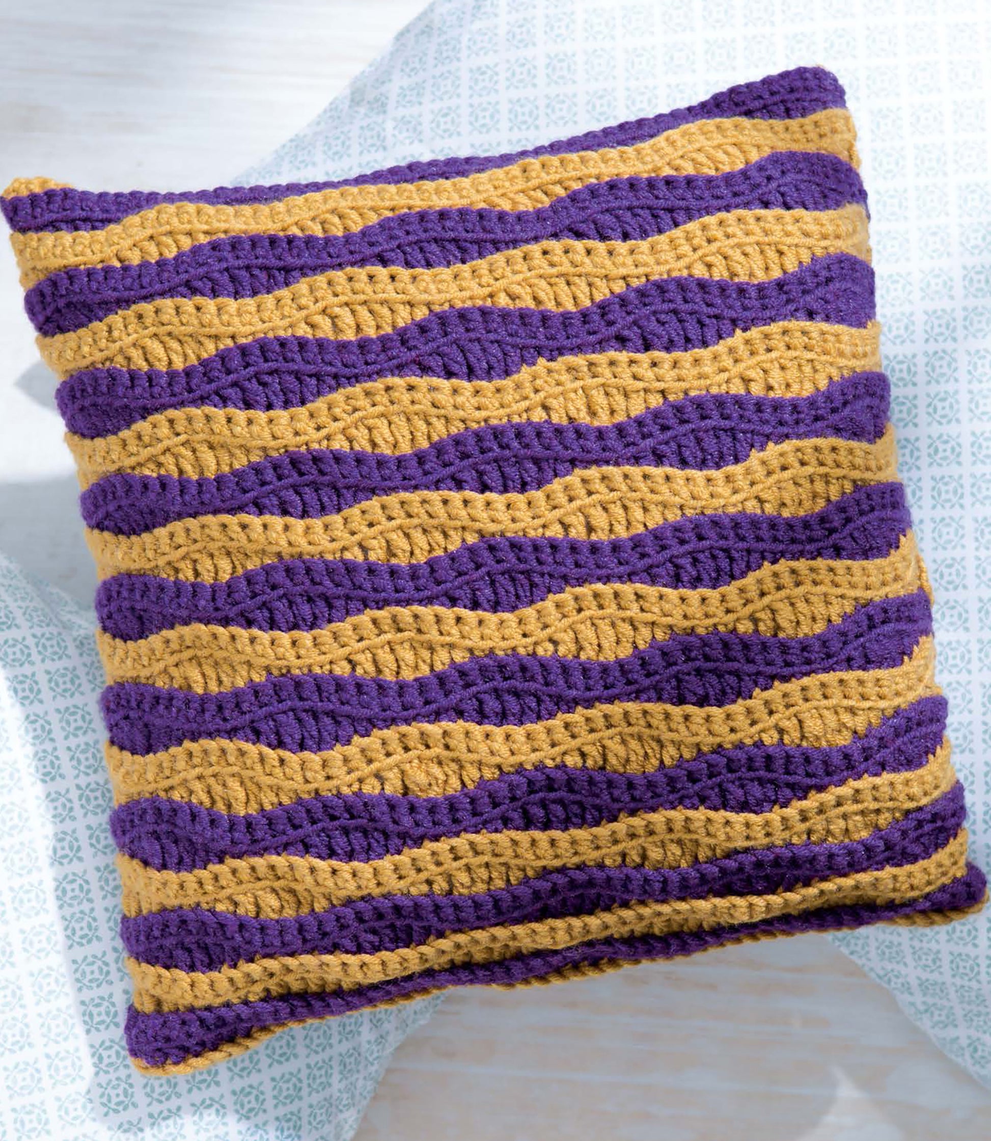 Textured Pillows Crochet by Leisure Arts features a square cushion with wavy purple and mustard yellow stripes on a light surface—ideal inspiration for unique crochet pillow patterns and textured crochet designs.