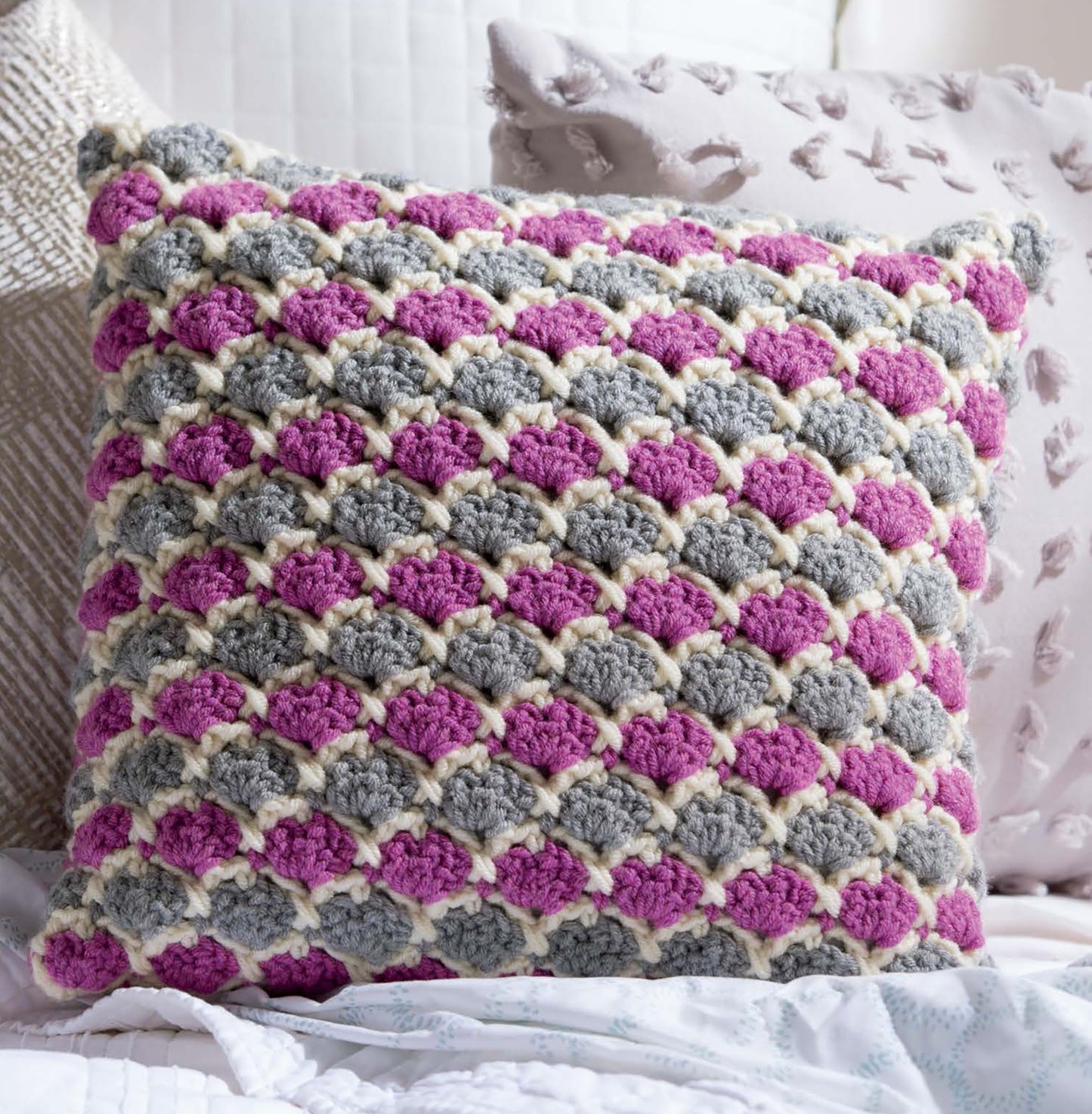 The Leisure Arts Textured Pillows Crochet features a square pillow with scalloped patterns in gray, pink, and cream—ideal for anyone seeking unique textured crochet designs—shown resting among neutral-toned decorative pillows.
