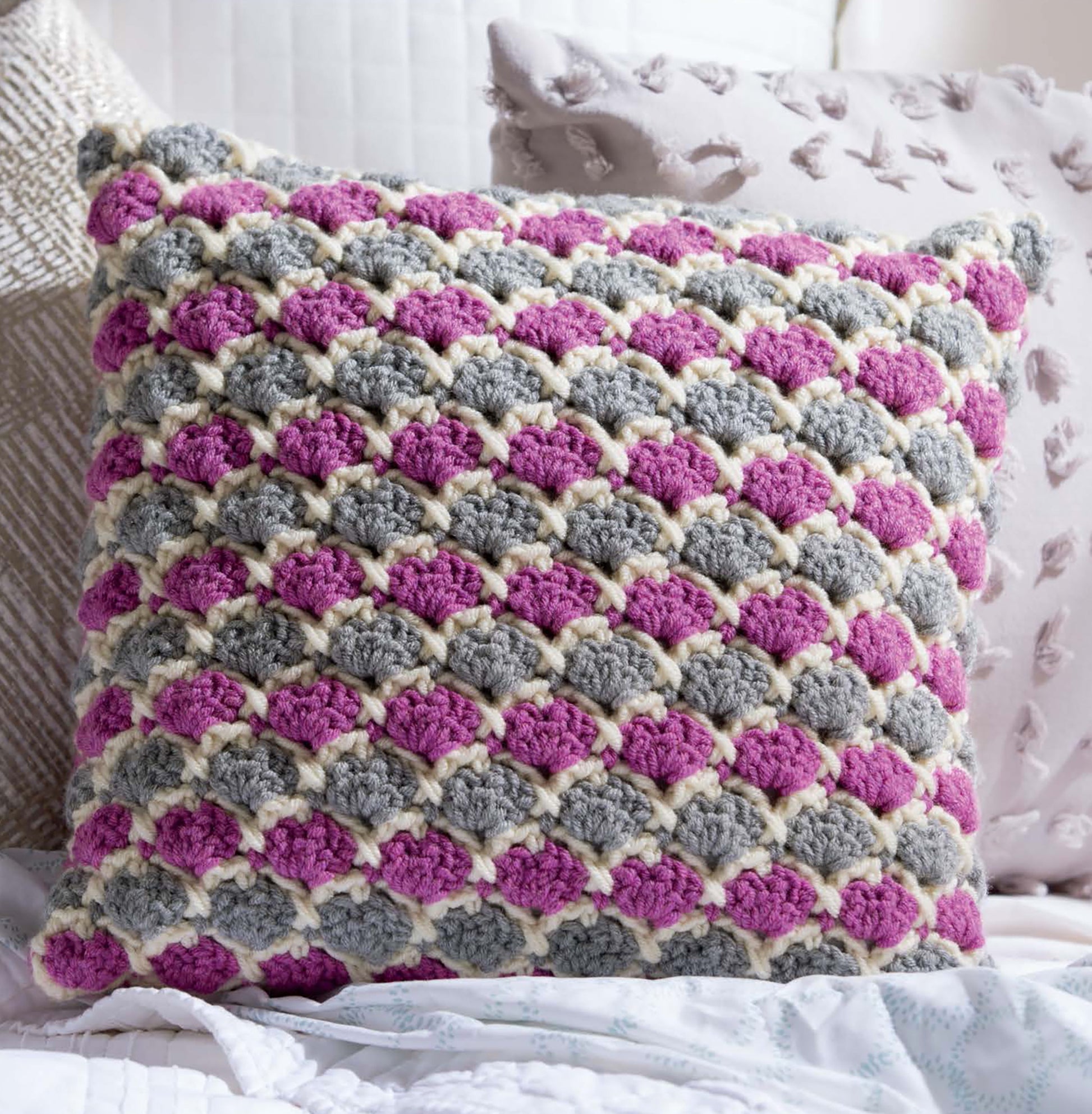 The Leisure Arts Textured Pillows Crochet features a square pillow with scalloped patterns in gray, pink, and cream—ideal for anyone seeking unique textured crochet designs—shown resting among neutral-toned decorative pillows.