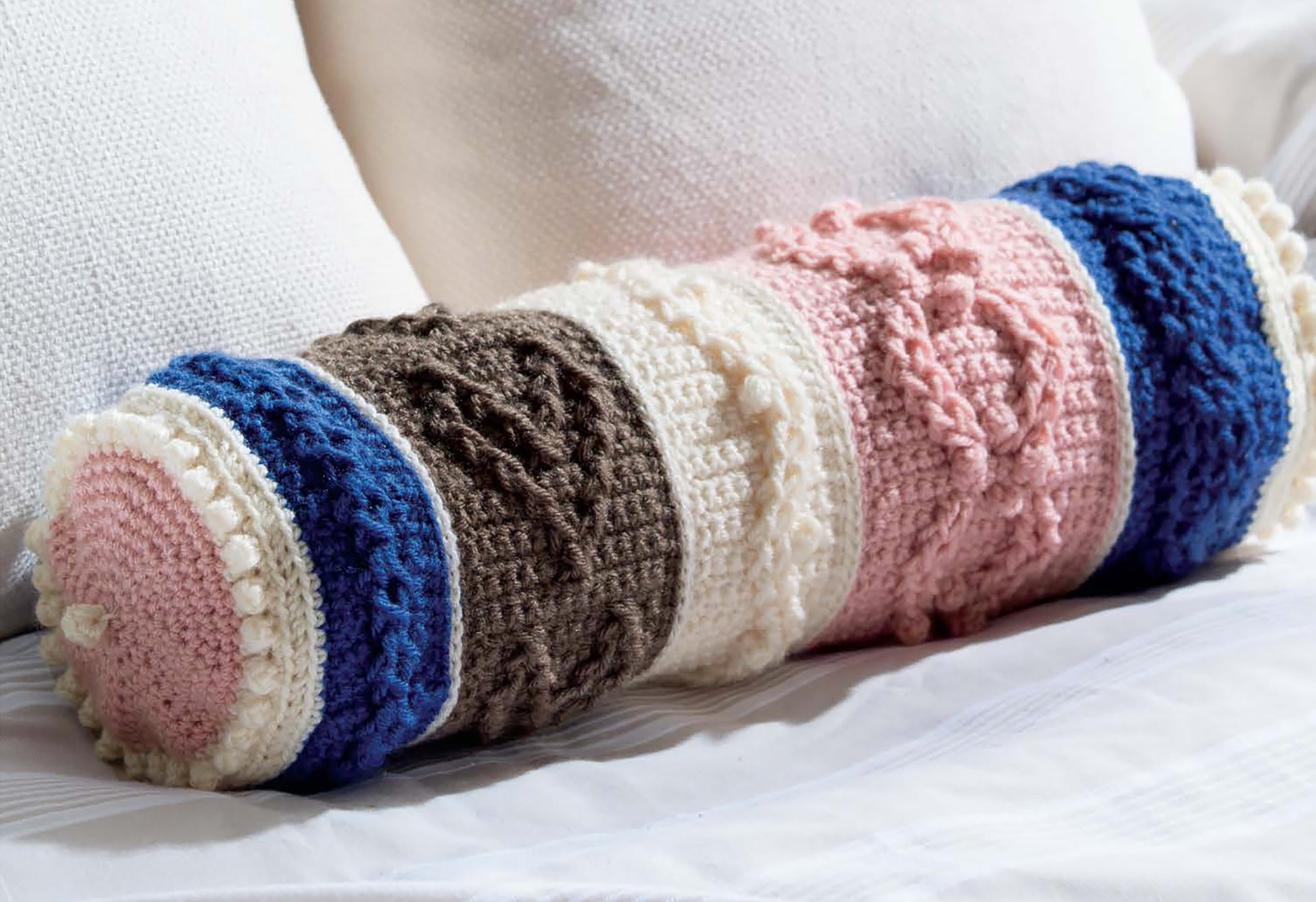 Textured Pillows Crochet by Leisure Arts features a cylindrical knit pillow with blue, brown, cream, and pink stripes—an ideal source of inspiration for anyone looking for new crochet pillow patterns or unique textured designs.