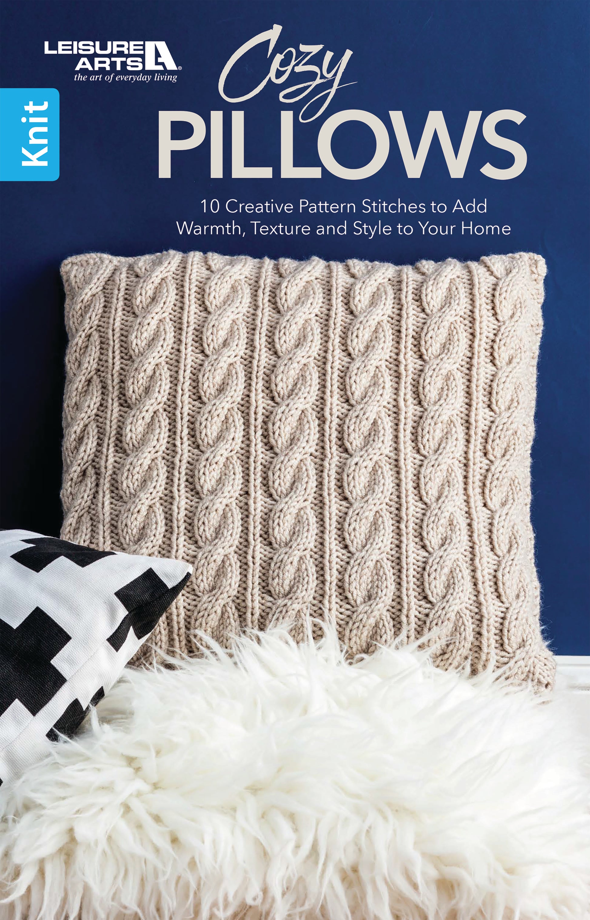 A soft beige cable-knit pillow rests on a white faux fur throw beside a black and white geometric pillow, set against a deep blue wall. Cozy Pillows by Leisure Arts: 10 creative knitting stitches to add warmth, texture, and style to your home.