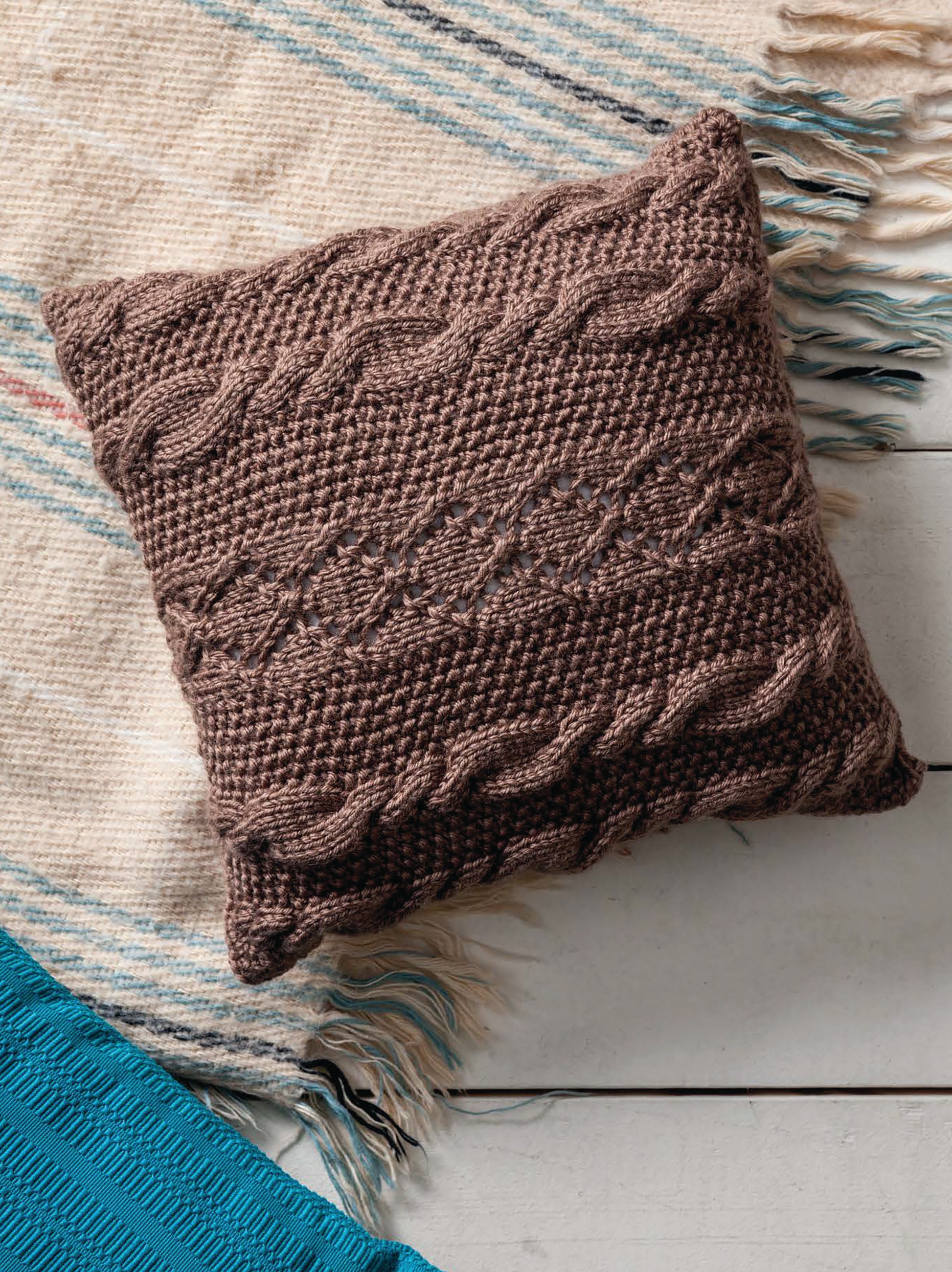 The Cozy Pillows by Leisure Arts—a brown, cable-patterned knitted pillow—rests on a cream fringed rug with blue stripes beside a blue textile atop a white wooden floor.