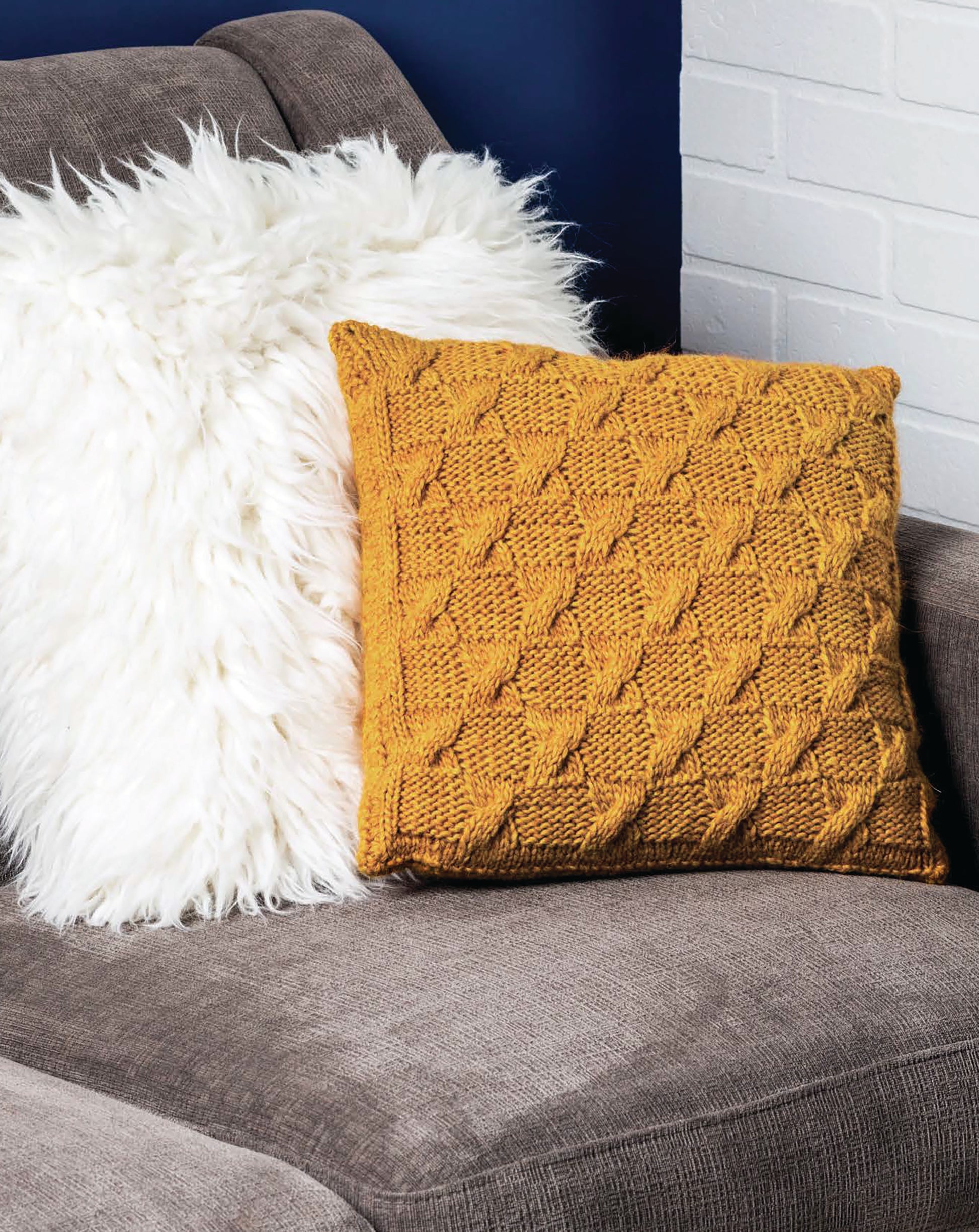 The Cozy Pillows by Leisure Arts features a mustard yellow pillow with a creative knitted texture on a gray sofa beside a fluffy white pillow, set against blue and white brick walls.