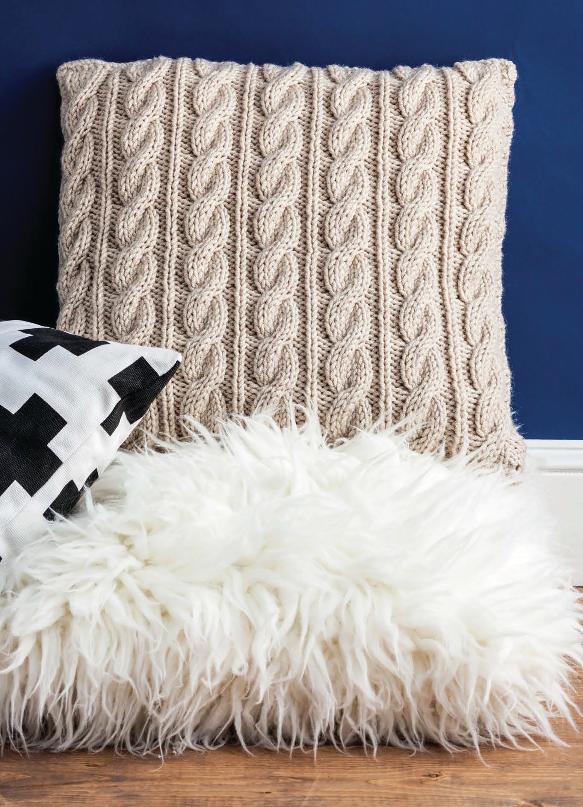 A cozy scene with Leisure Arts’ Cozy Pillows—beige cable-knit and black-and-white patterns—paired with a fluffy white rug, pops against a dark blue wall and wood floor, highlighting stylish knit home decor.