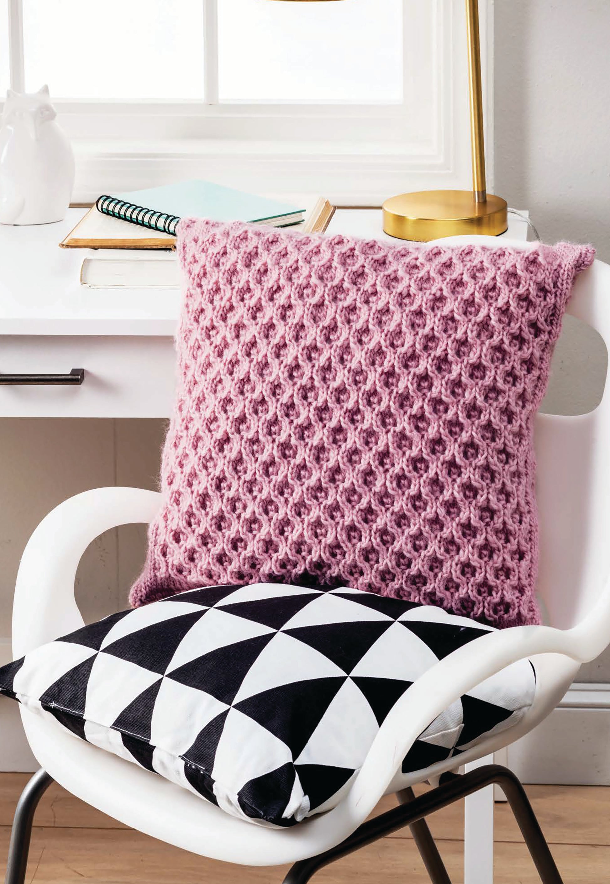 A white chair holds two Cozy Pillows from Leisure Arts—one with a pink knitted cover, the other in a black-and-white geometric print. Behind the chair sits a white desk with books, a lamp, and a ceramic cat.