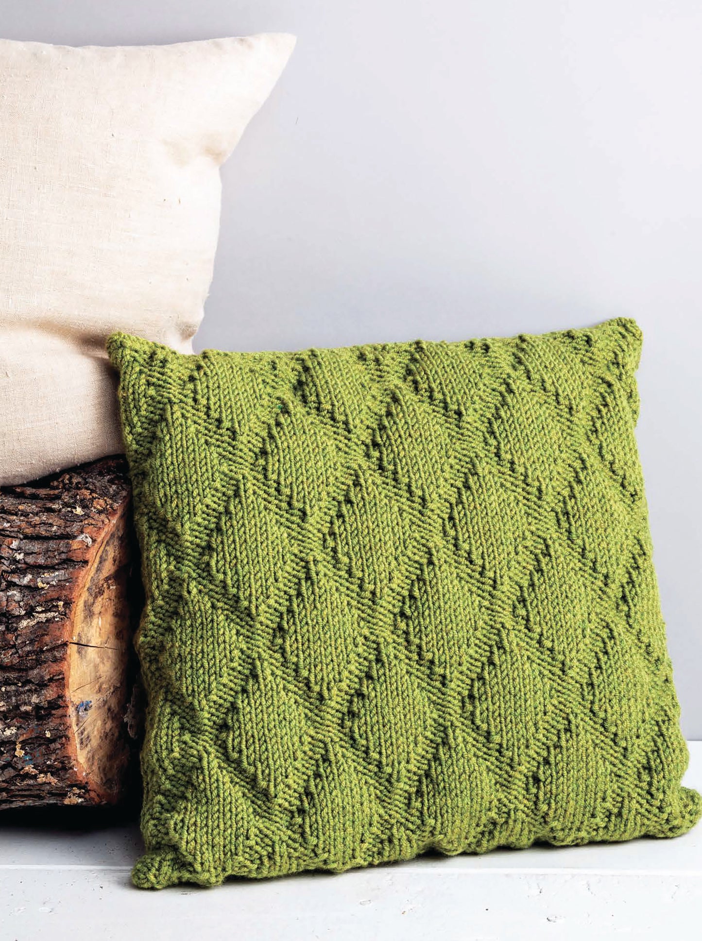 The Cozy Pillows by Leisure Arts, featuring creative green pillow knitting with a diamond pattern, rests against a light beige pillow and a log against a plain light gray background.