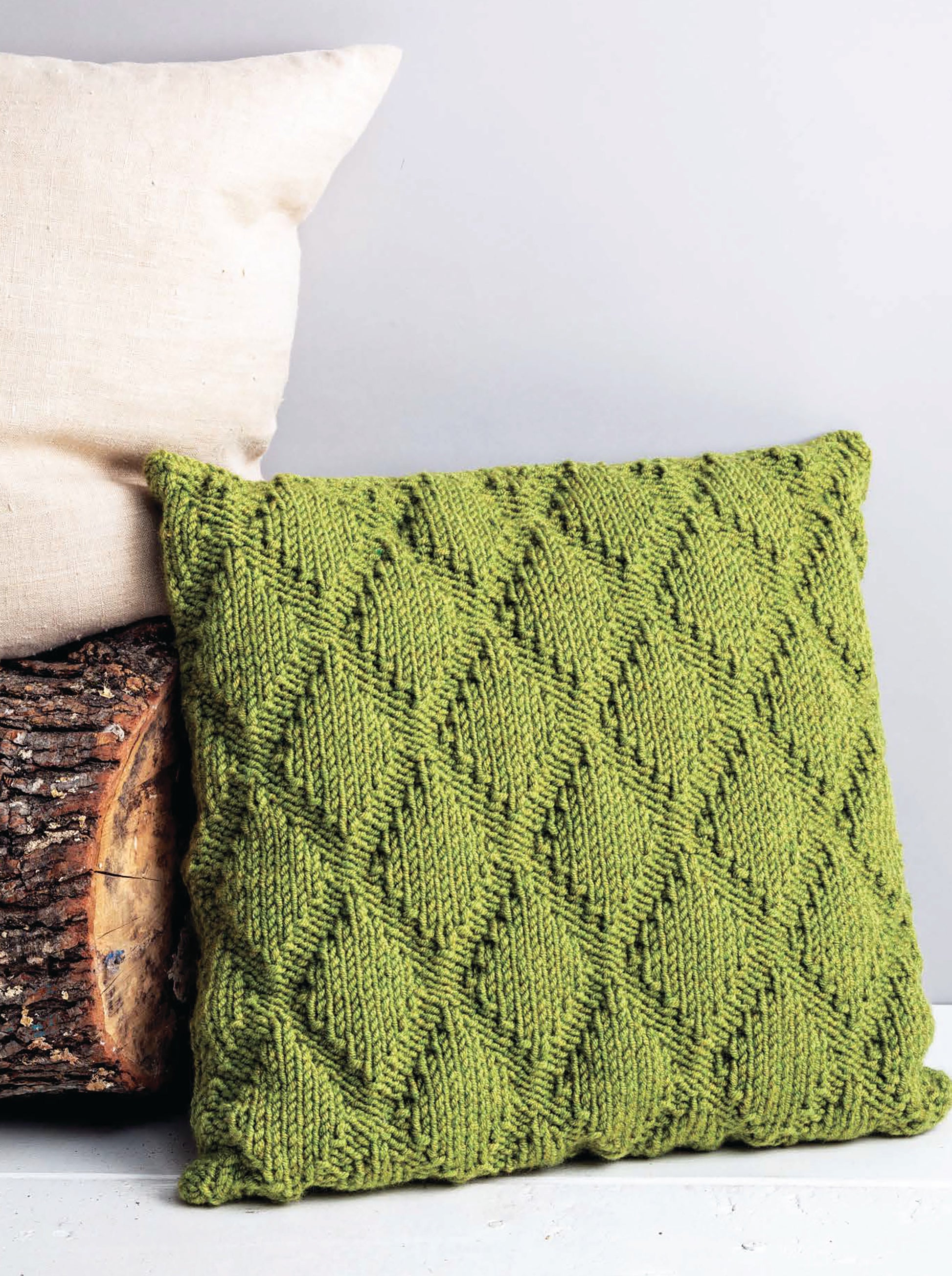 The Cozy Pillows by Leisure Arts, featuring creative green pillow knitting with a diamond pattern, rests against a light beige pillow and a log against a plain light gray background.