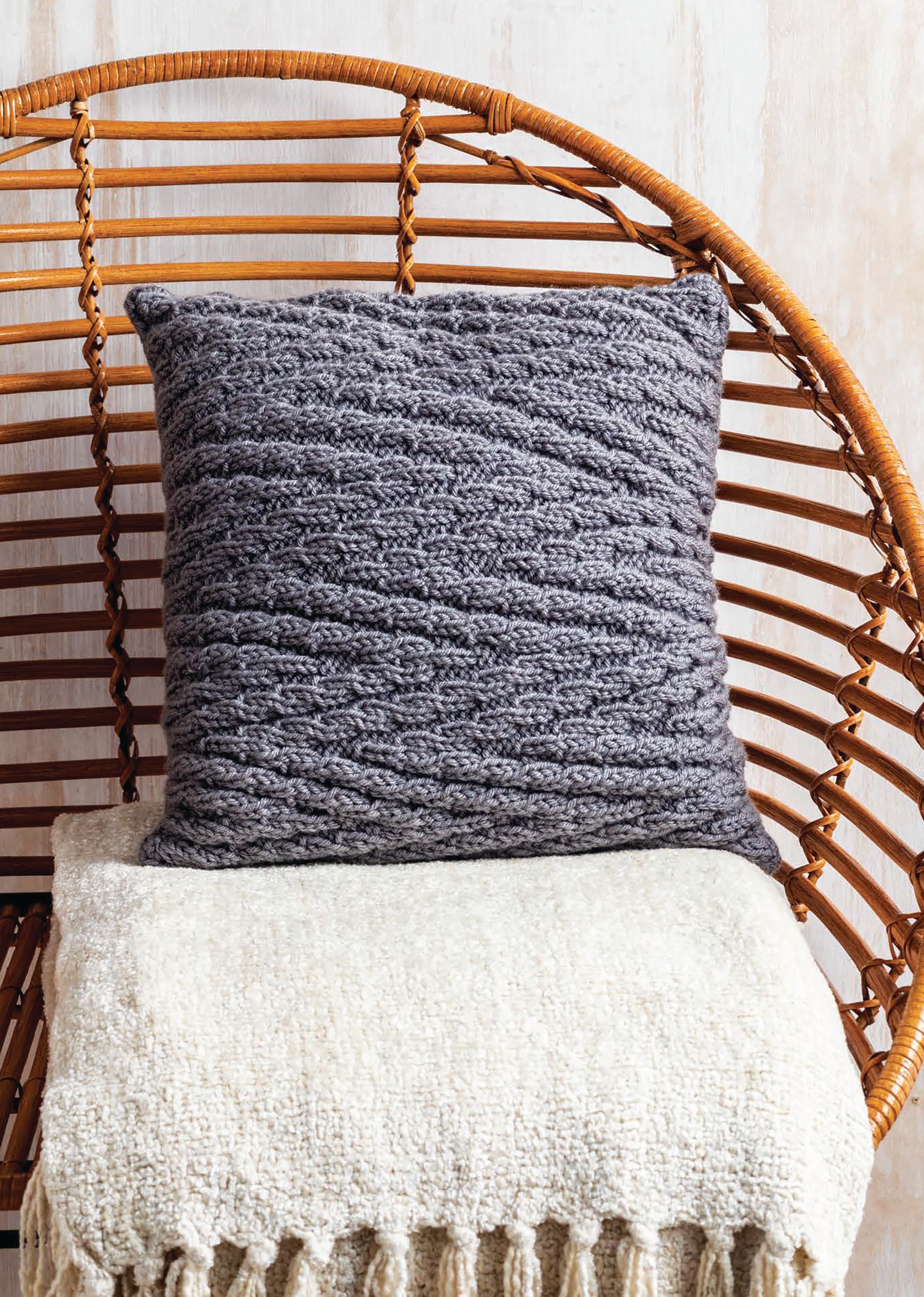 The Leisure Arts Cozy Pillows, featuring creative textured knitting in gray, sit on a cream fringed blanket atop a curved wicker chair against a light wall.