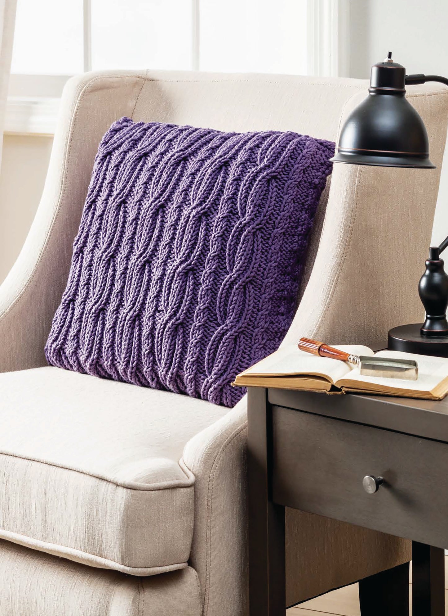 A beige armchair with a Leisure Arts Cozy Pillows textured purple pillow is beside a dark wood table holding an open book, glasses, and a black lamp, all near a window with light curtains.