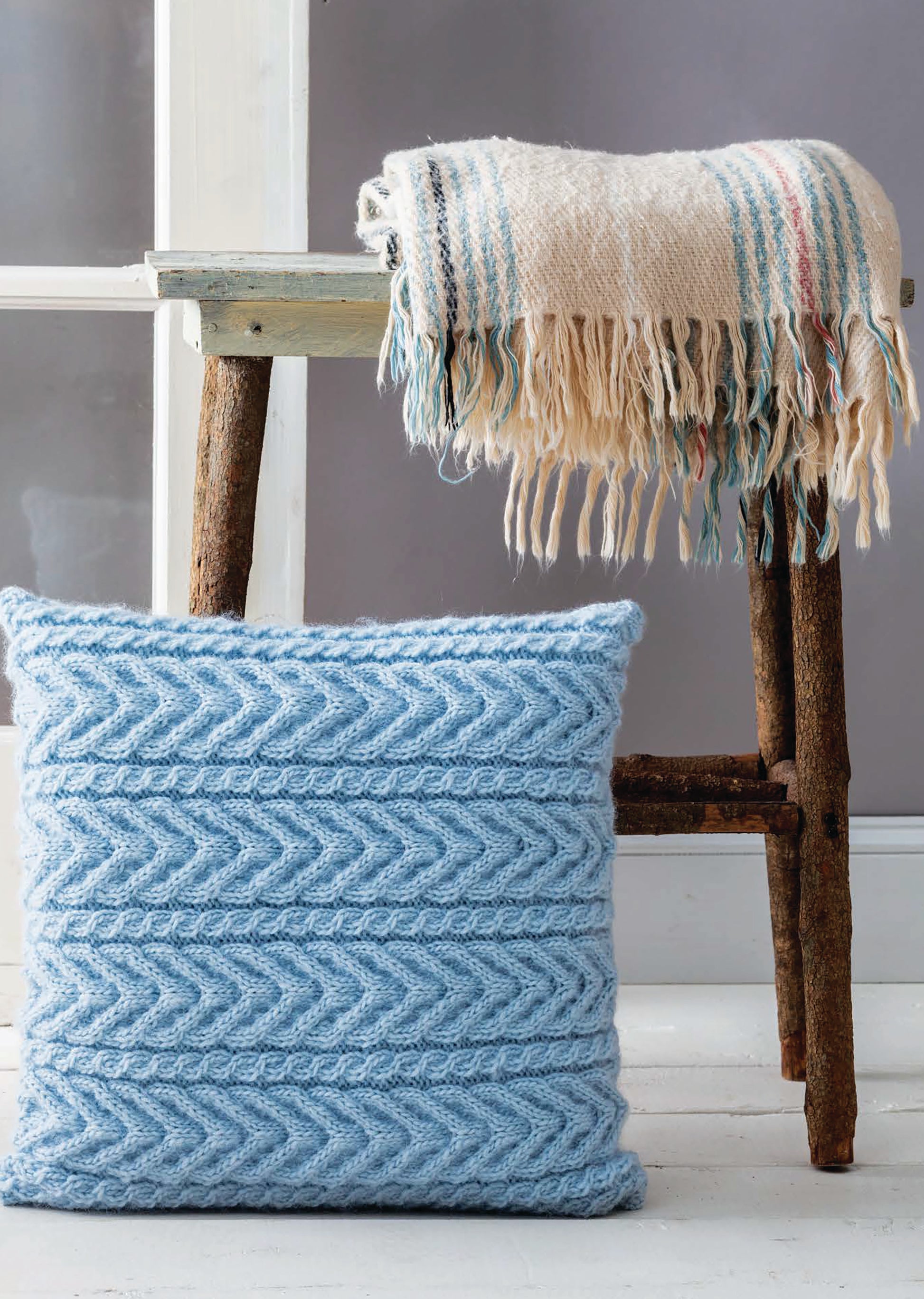 The Cozy Pillows by Leisure Arts features a light blue knitted chevron-pattern pillow beside a rustic wooden stool, topped with a cream fringed blanket featuring blue and red stripes.