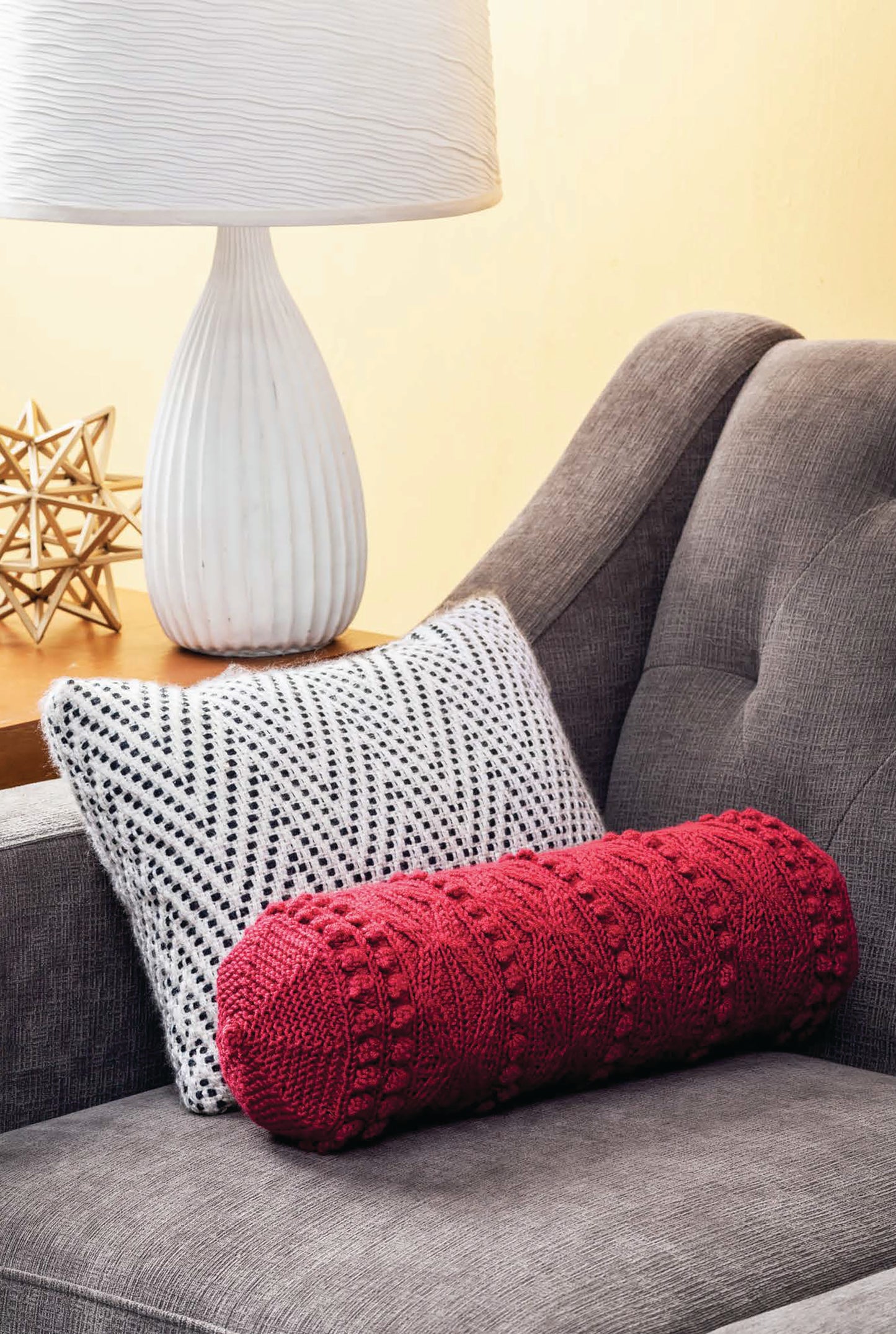 The Cozy Pillows Digital Download by Leisure Arts features a knitted red bolster pillow on a gray sofa with a black-and-white patterned pillow, while a white lamp and gold geometric decor accent the side table in the background.