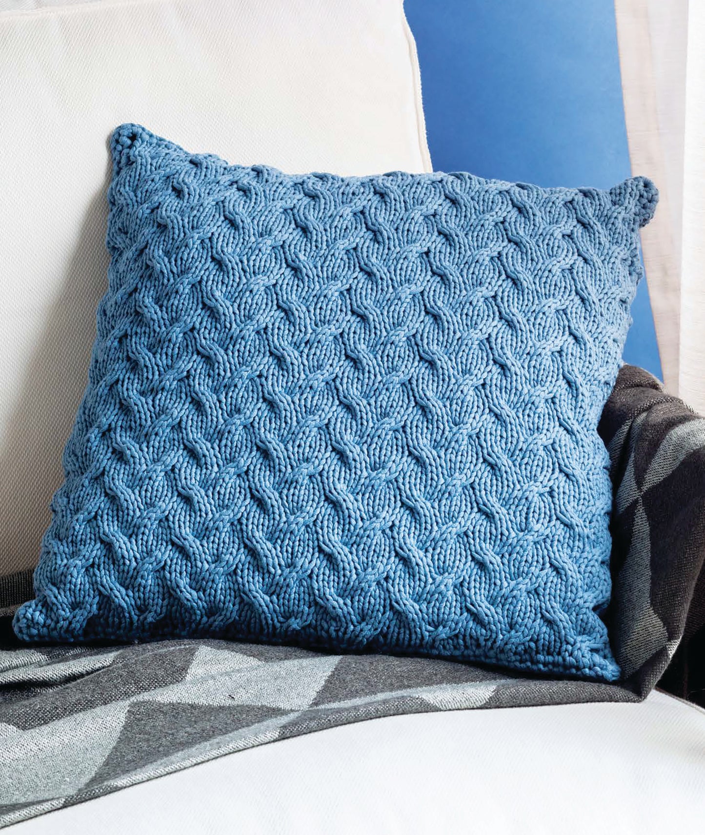 The Leisure Arts Cozy Pillows, a blue, textured knitted throw with a cable pattern, sits on a white couch beside a gray and black geometric blanket.