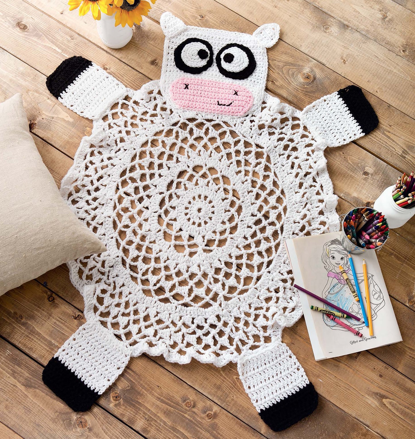 The Leisure Arts Crochet Rugs for Kids features a cow-shaped crochet rug with a lacy white body and black-tipped legs—ideal playroom decor or a charming children’s gift alongside pillows, colored pencils, coloring books, or yellow flowers.