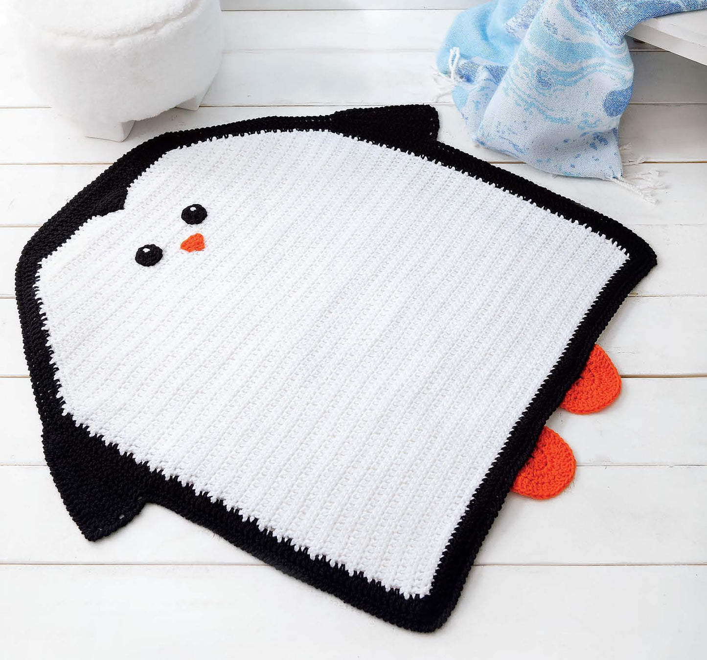 The Leisure Arts Crochet Rugs for Kids, featuring a penguin design with a white body, black wings and trim, orange beak and feet, and black eyes, adds playful charm to any playroom decor. Shown here on a white wooden floor near a blue blanket and ottoman.