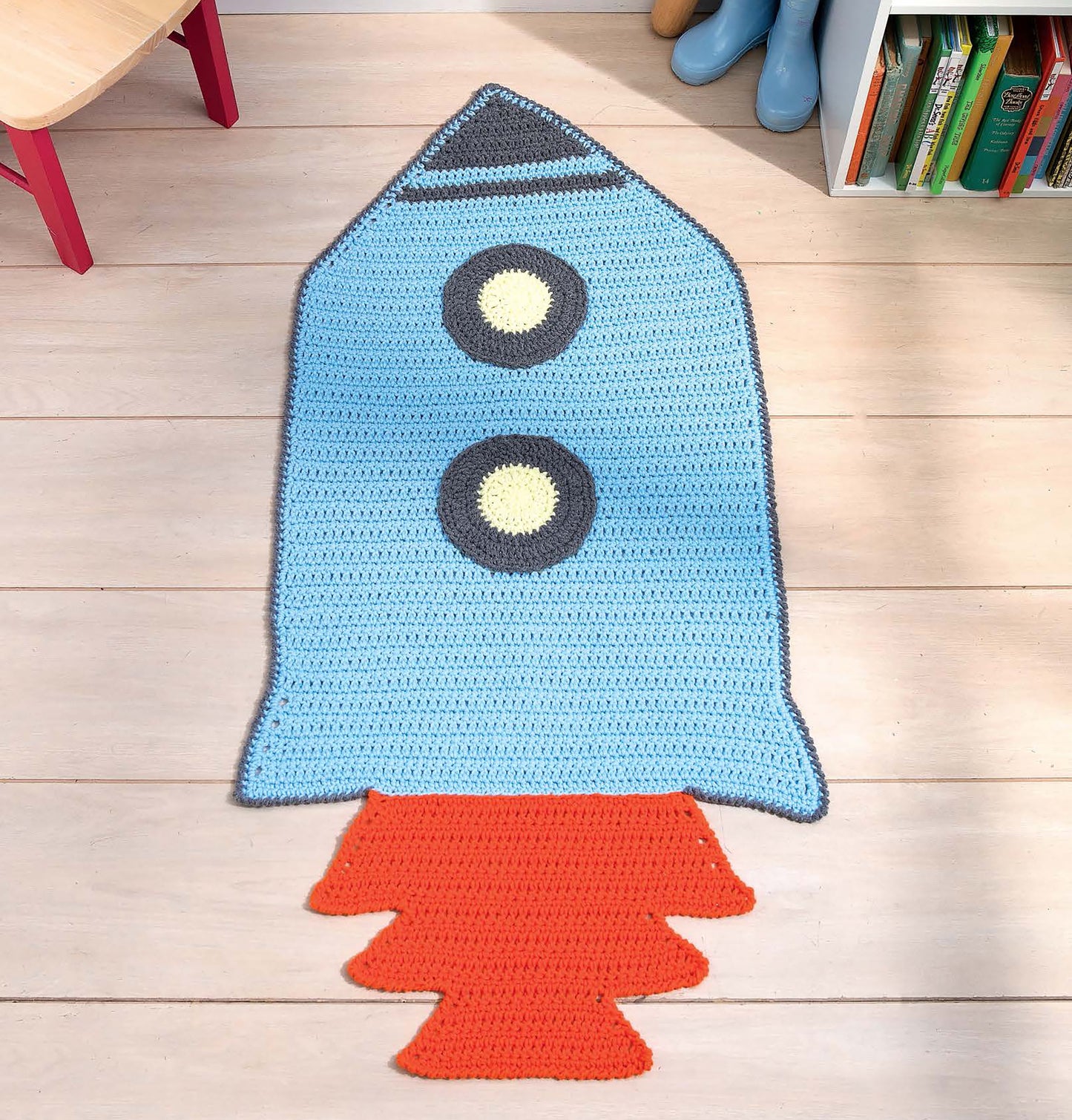 The "Crochet Rugs for Kids" by Leisure Arts is a blue rocket-shaped crocheted rug with yellow windows and an orange flame, perfect for playful playrooms or as a unique gift for children. Shown on a wooden floor near a bookshelf and blue boots.