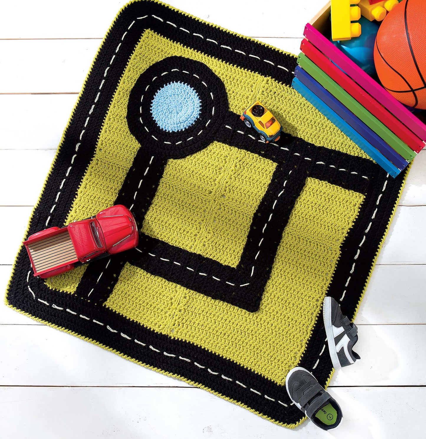 The Leisure Arts Crochet Rugs for Kids features a yellow and black road-themed play mat, accessorized with toy cars, a basketball, books, and shoes on a white wooden floor—ideal for beginner projects or creative playroom decor.