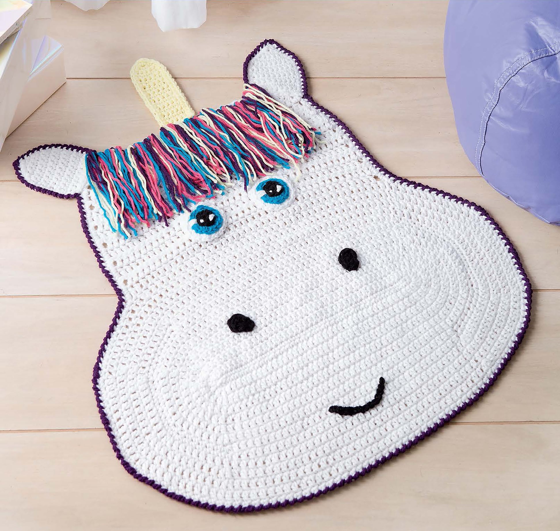Leisure Arts Crochet Rugs for Kids: A unicorn face rug with a white base, blue eyes, black details, purple-edged ears, and a colorful yarn mane—ideal for children's gifts or playroom decor. Shown on a light wooden floor.