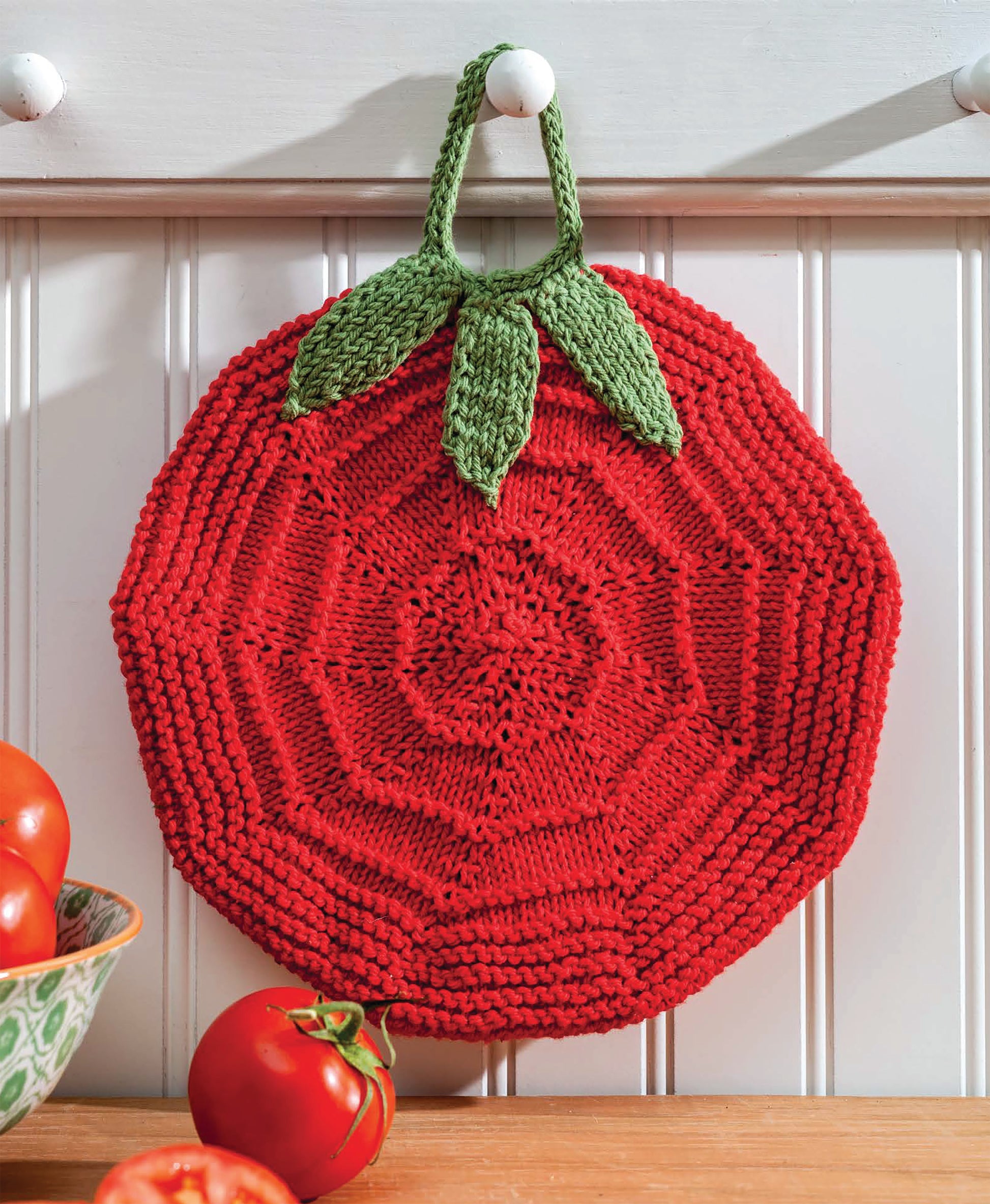 Displayed on a hook against a white paneled wall, the tomato-shaped potholder from Leisure Arts' "Make in a Weekend Potholders and Dishcloths" adds charm to any kitchen. Fresh tomatoes rest on the counter below.